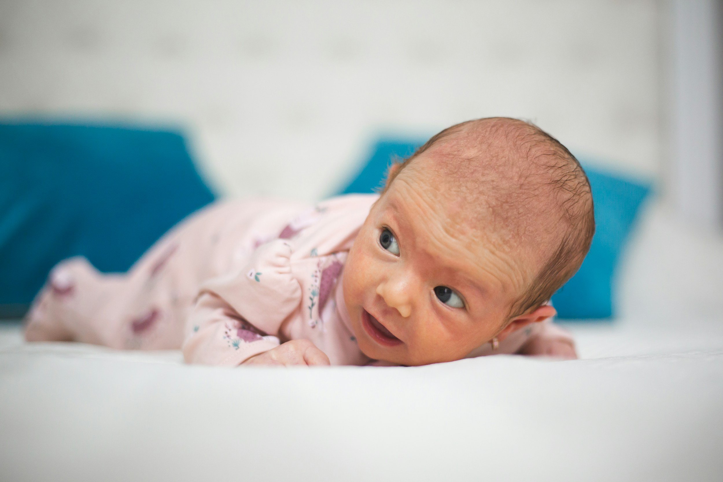 A baby girl lying on her stomach on a bed, looking to the side with wide eyes.