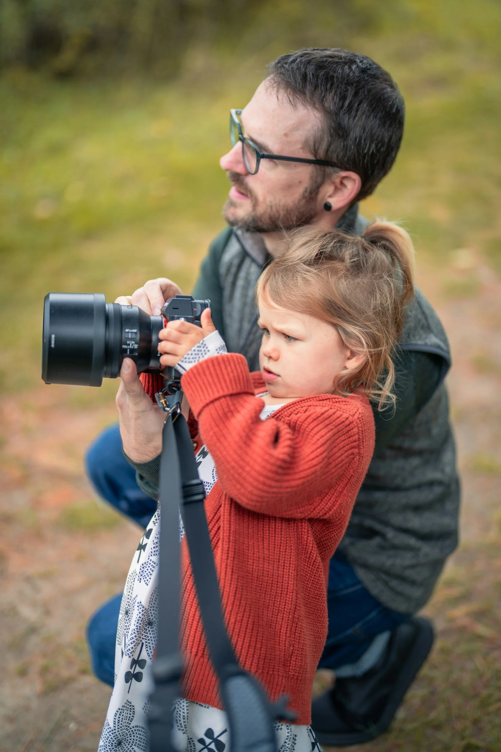 A man and a young girl outdoors, with the girl holding a camera, looking focused on taking a picture.