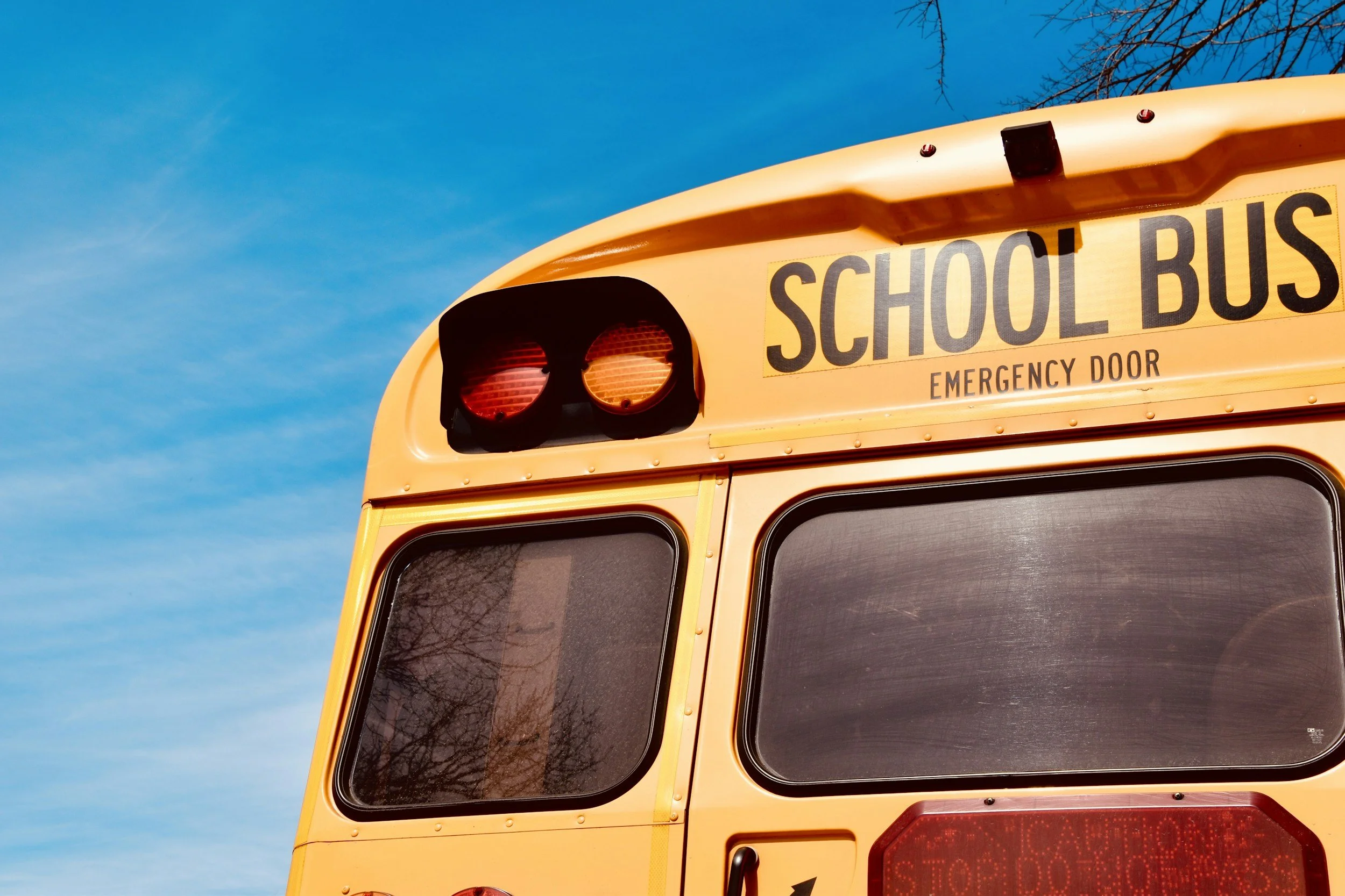Close-up of the front of a yellow school bus with red warning lights on, an emergency door sign, and tinted windows. Clear blue sky in the background.