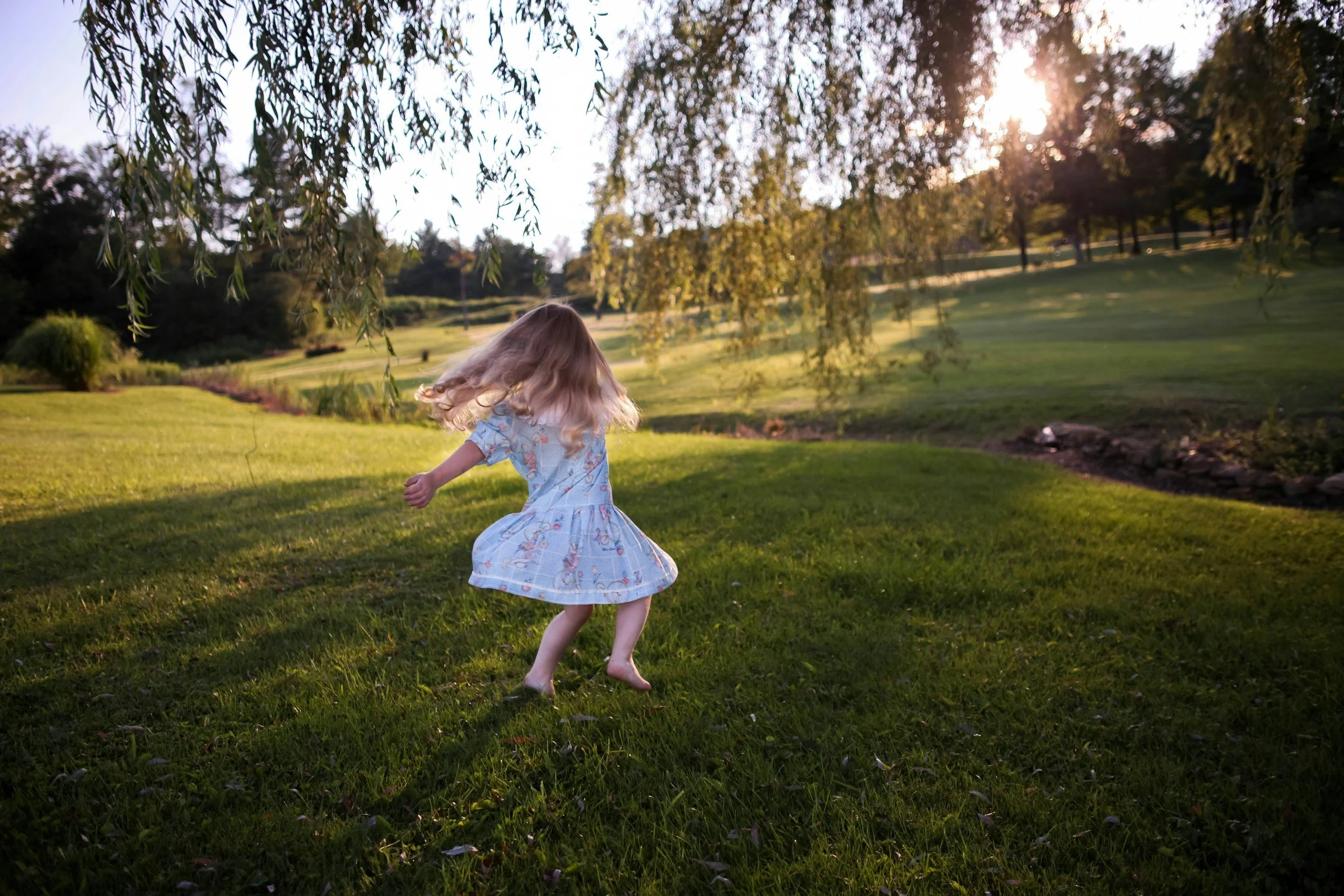 A young girl spinning and dancing on a grassy field at sunset, wearing a light blue dress with pastel patterns, with trees and a creek in the background.