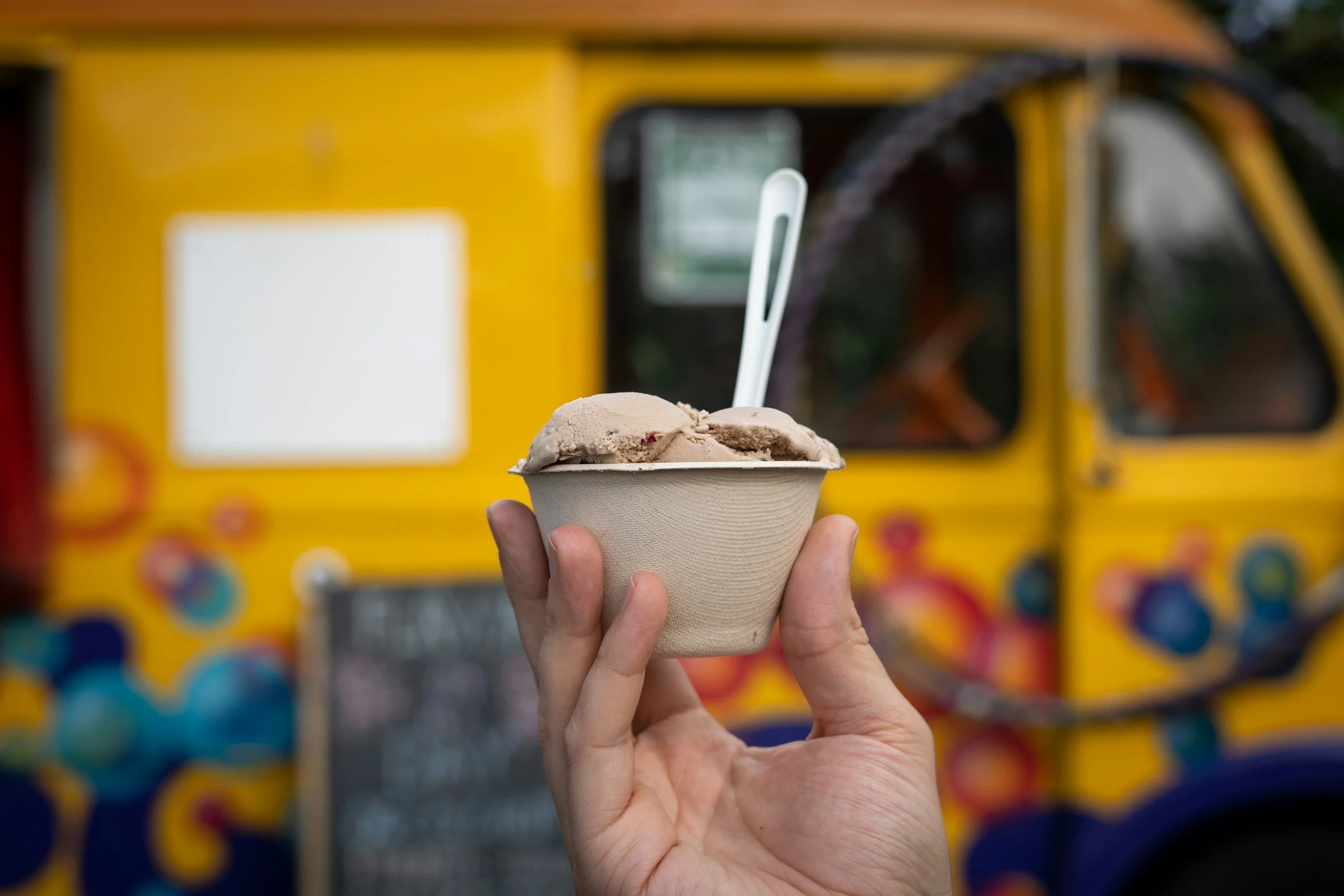 A hand holding a cup of ice cream with a spoon, with a colorful food truck in the background.