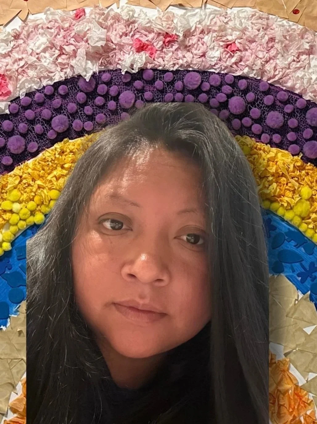 A woman with long dark hair and a neutral expression, standing in front of a colorful rainbow made from various textured and colored paper and materials, including green at the top, followed by light blue, brown, white with pink accents, purple, yellow, and blue.