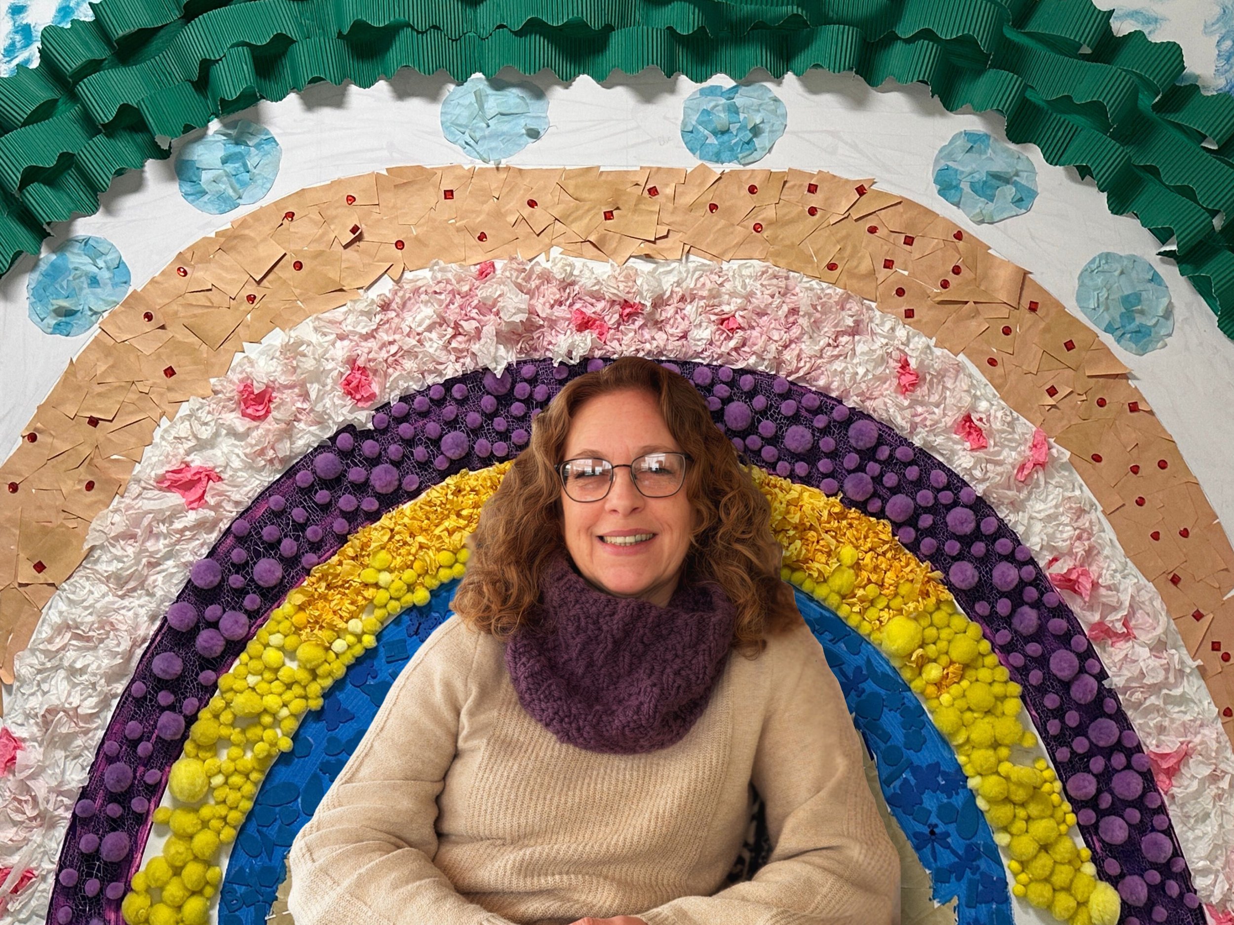 A woman with curly hair and glasses smiling while sitting in front of a colorful rainbow made of various textured craft materials.