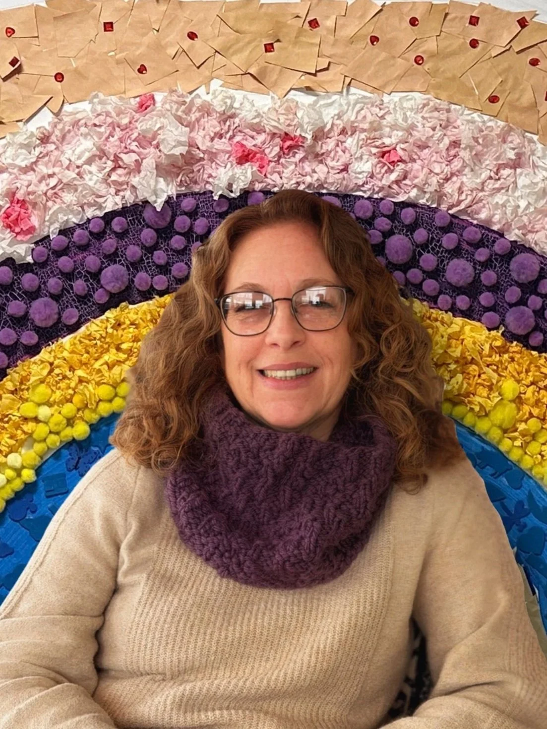 A woman with curly hair and glasses smiling while sitting in front of a colorful rainbow made of various textured craft materials.