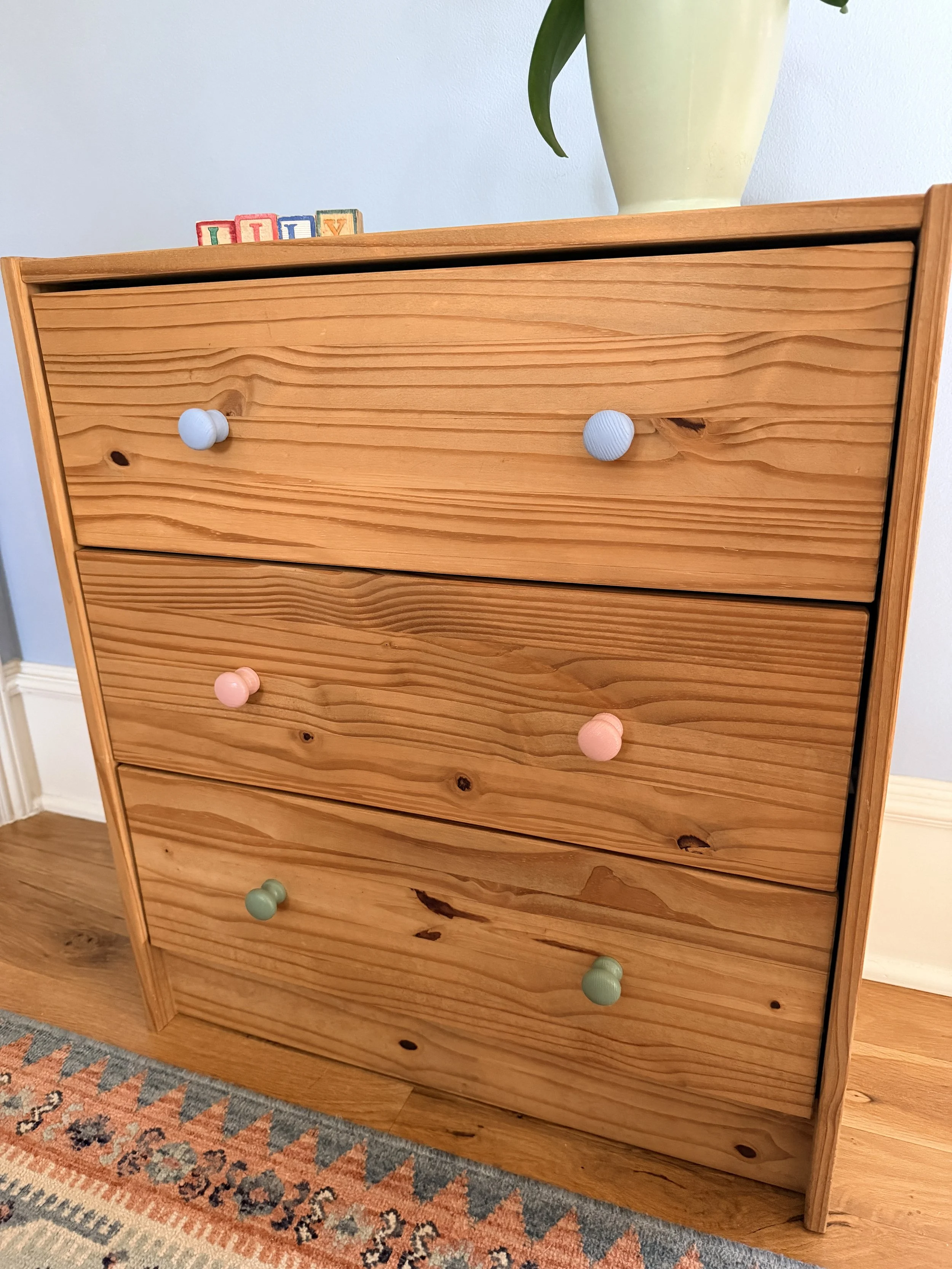 A wooden dresser with four drawers, each with colorful round knobs, placed on a hardwood floor near a patterned carpet, with a vase with a plant on top.