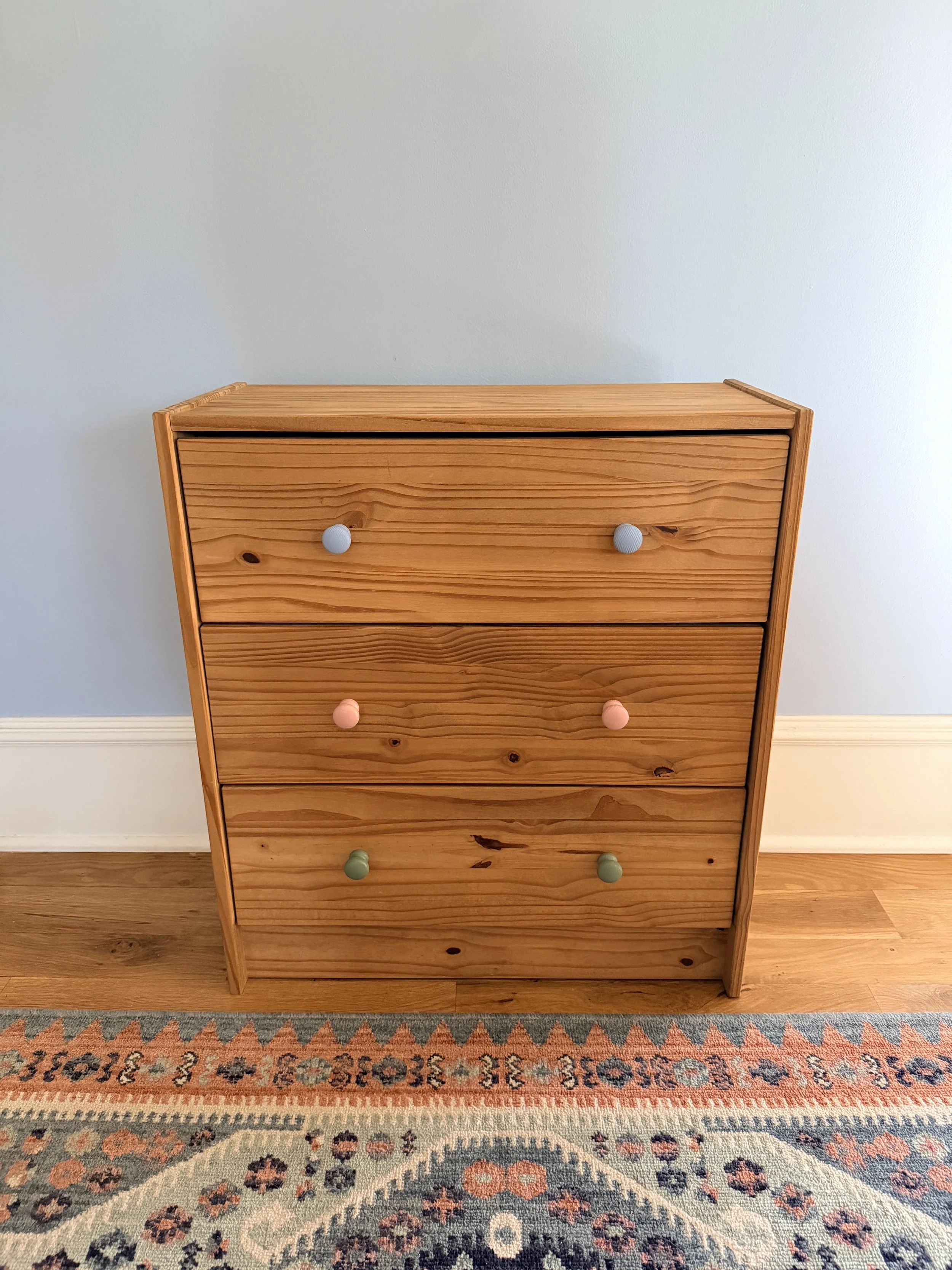 A wooden dresser with four drawers, each with painted knobs in different pastel colors, against a light blue wall and placed on a patterned rug.