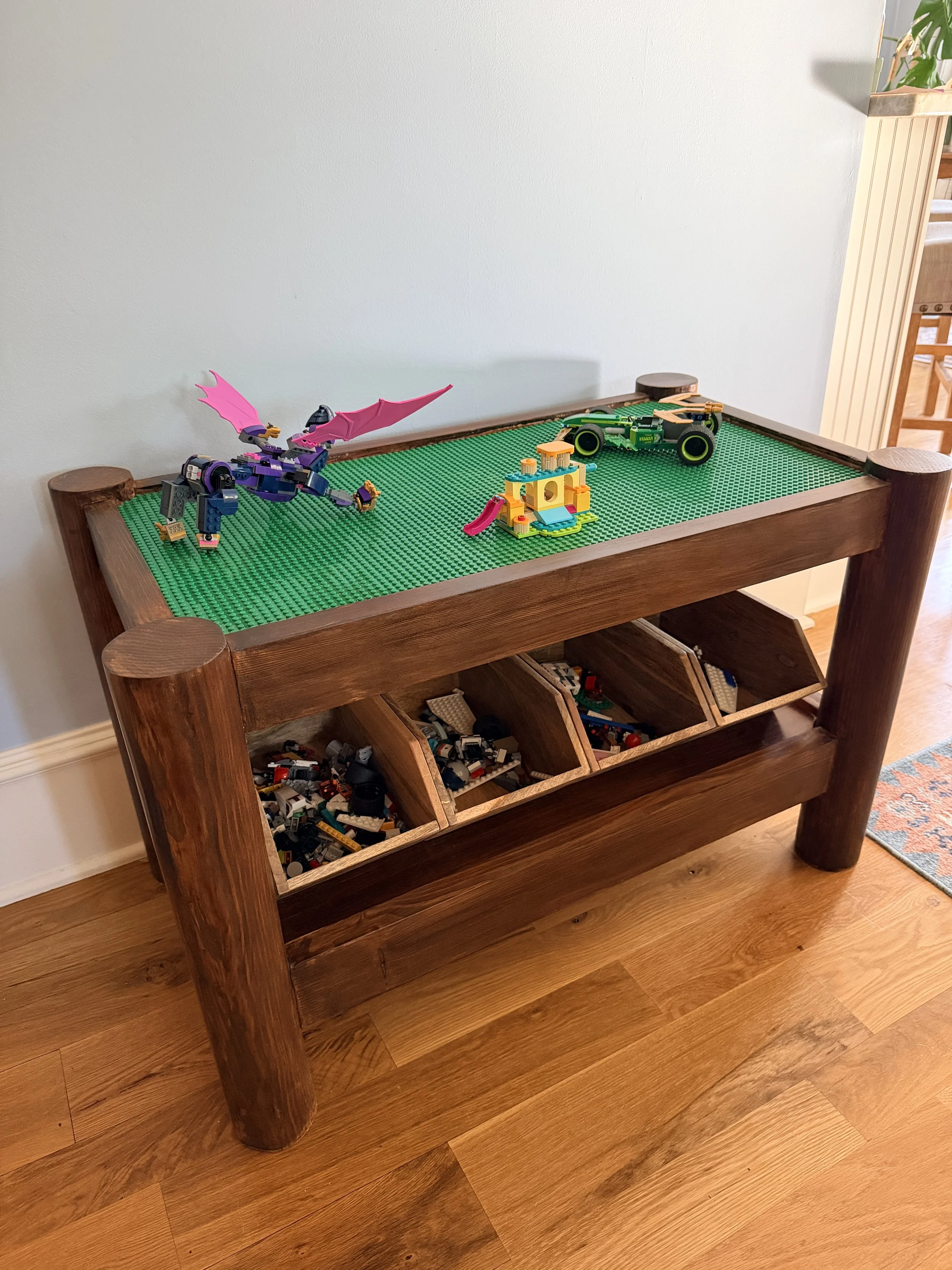 A wooden table with lego creations on top, including a purple dragon, a green race car, and a small colorful house, with labeled compartments filled with additional lego pieces below.