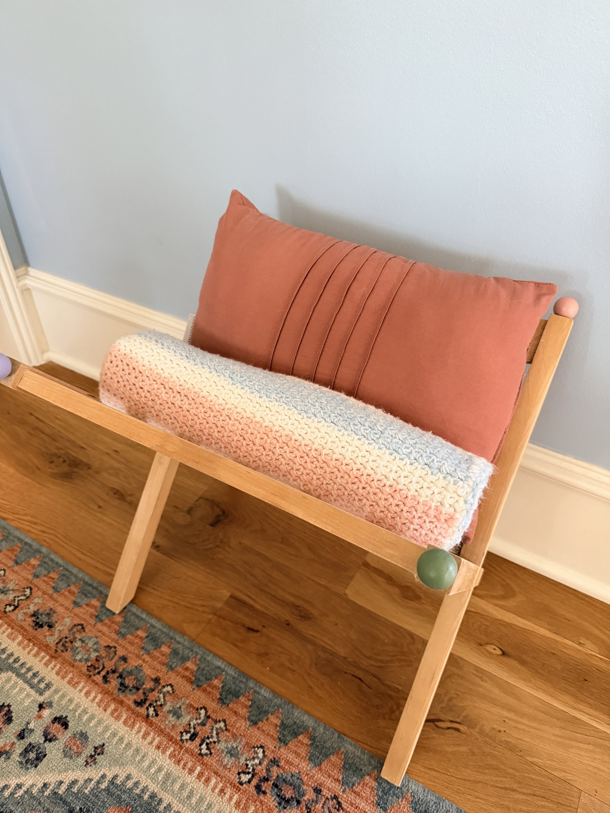 A wooden child-sized bench with a pink pillow and a chunky knit blanket, placed against a light blue wall on a patterned rug and hardwood floor.