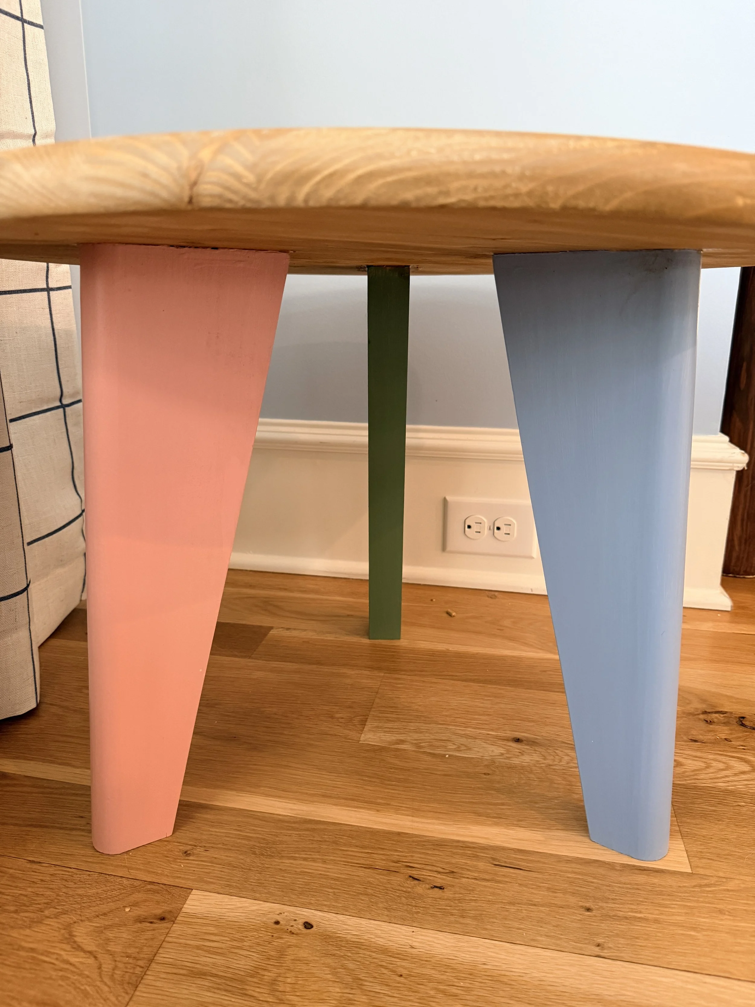 A close-up of a small, round wooden table with three angled legs painted in pastel colors: pink, green, and blue, on a hardwood floor against a light blue wall.