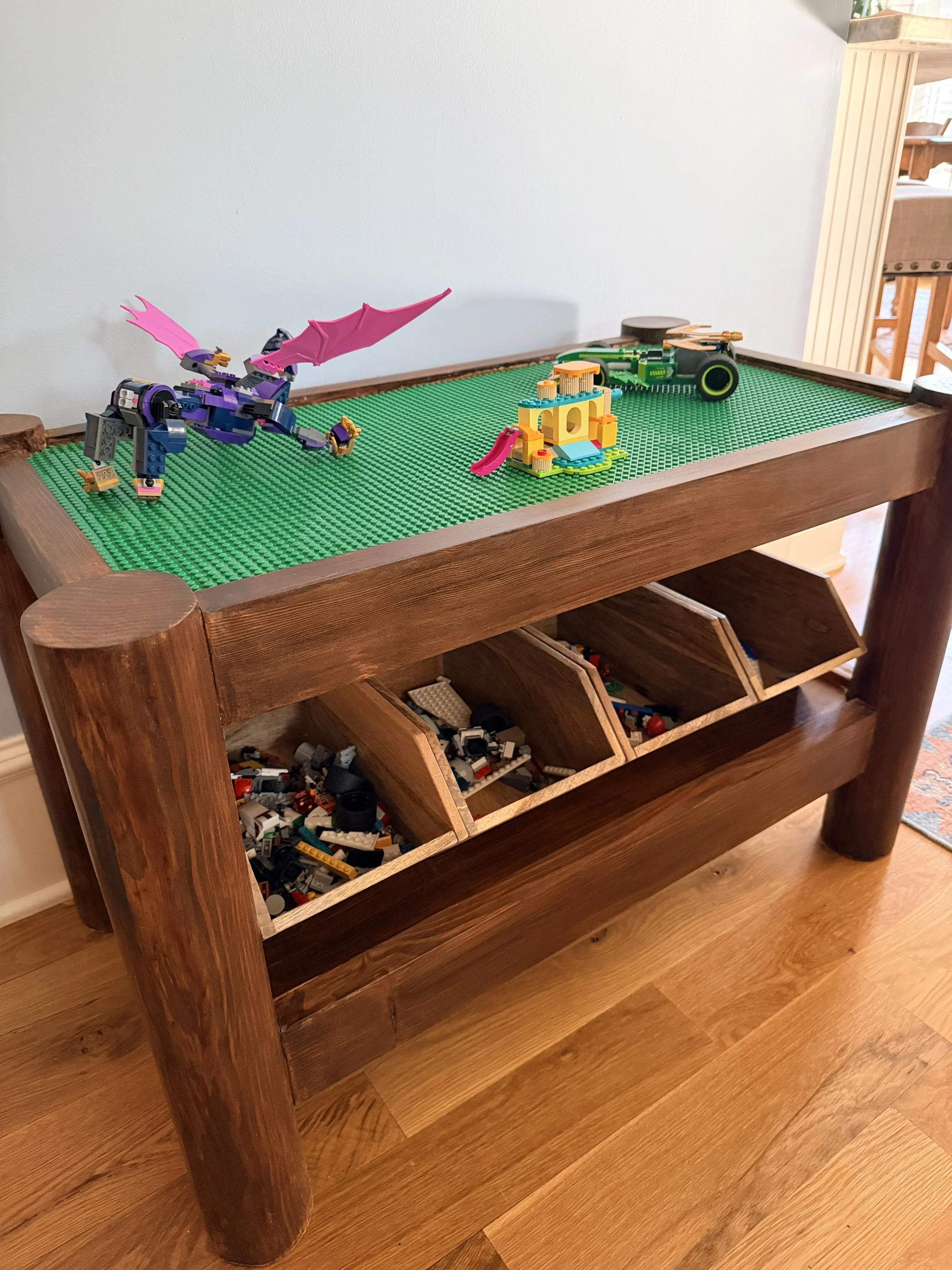 A wooden table with a green LEGO baseplate on top, displaying LEGO builds including a purple dragon, a yellow playhouse with a slide, and a green construction vehicle. The table includes open storage compartments filled with LEGO pieces.