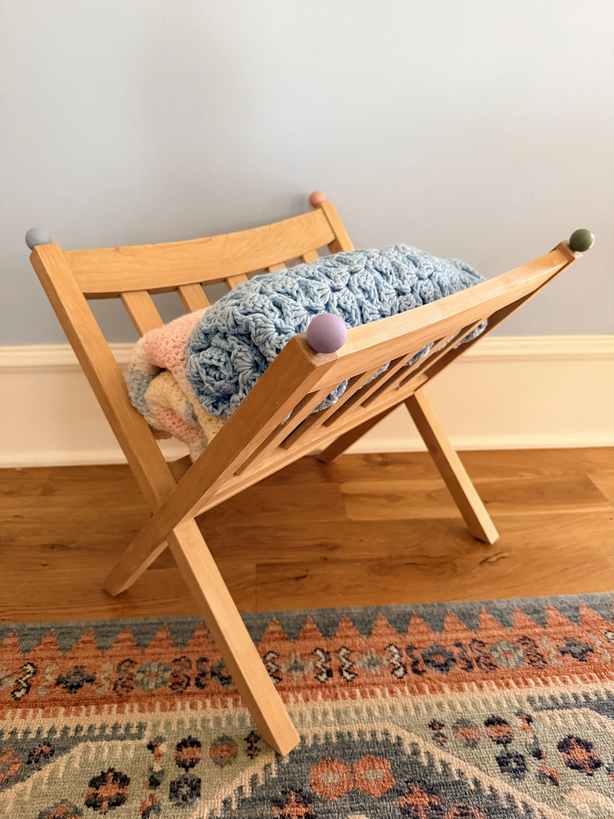 Wooden drying rack with colorful knobs at the ends, holding a crocheted blanket and a folded plush blanket, next to a patterned rug and a light gray wall.