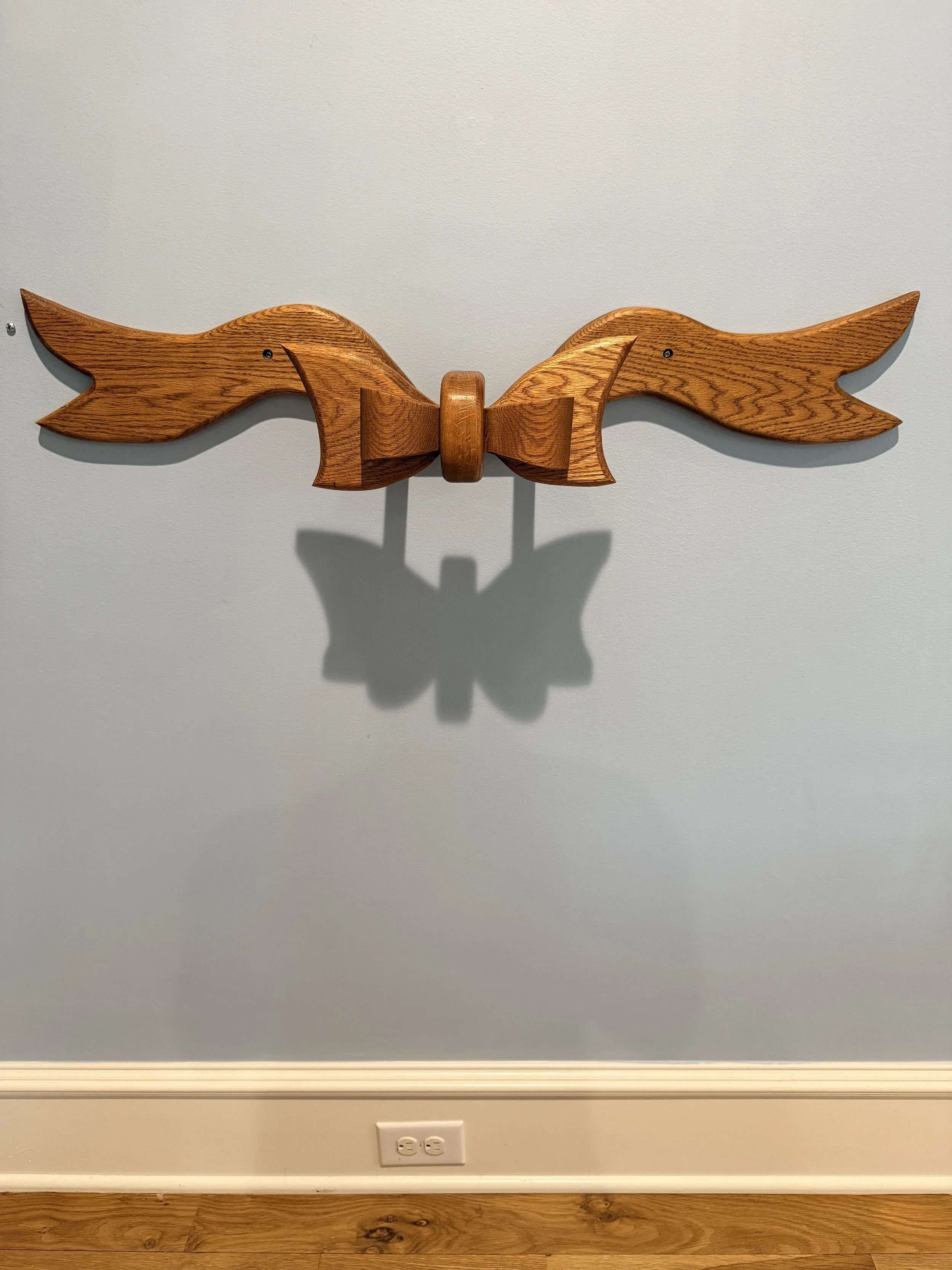 Wooden wall art in the shape of a bow tie with two winged shapes extending outwards, mounted on a light gray wall.