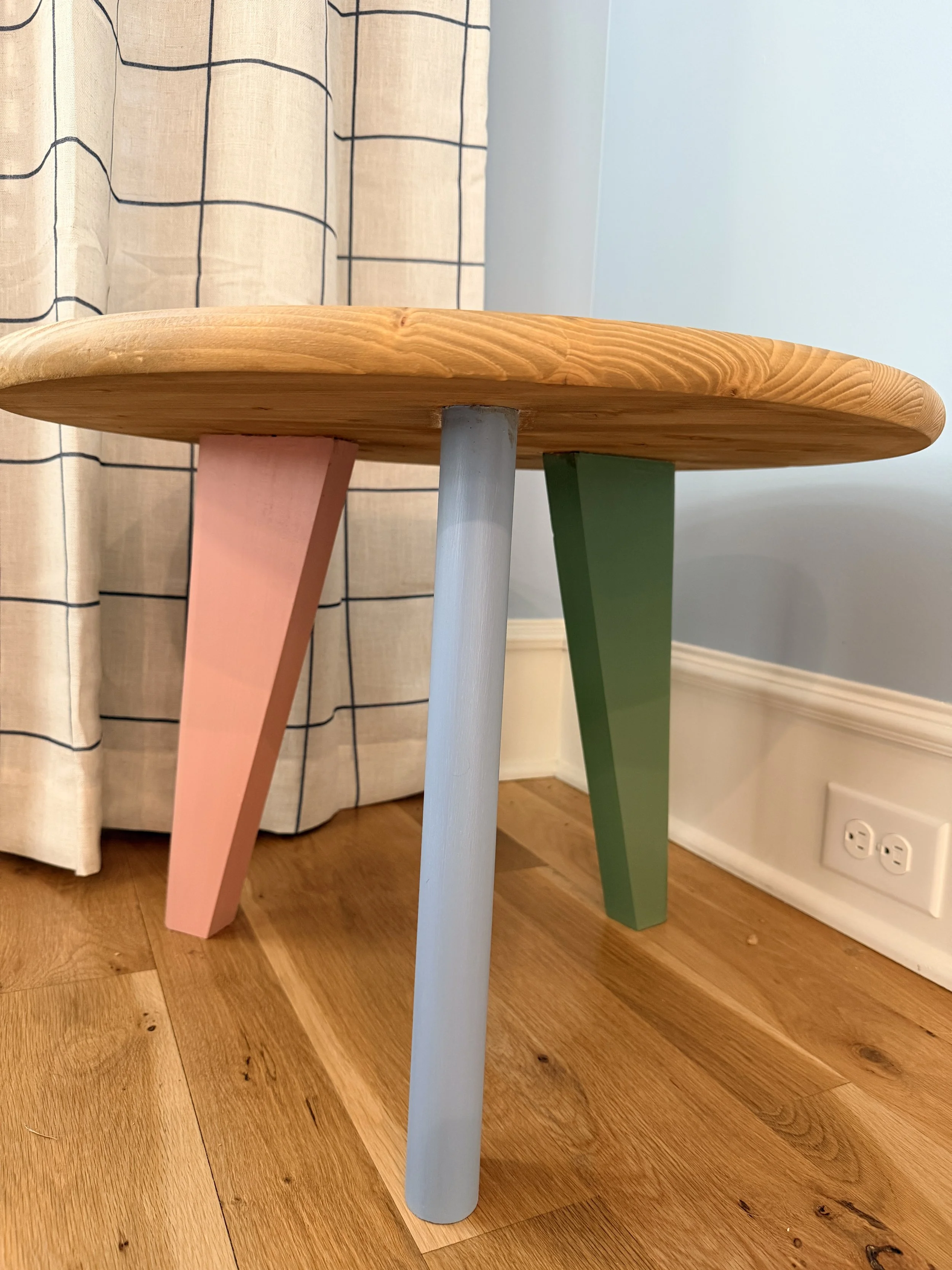 Colorful wooden stool with three angled legs painted in pink, white, and green, positioned on hardwood floor near a beige curtain with black grid lines and a white wall with electrical outlets.