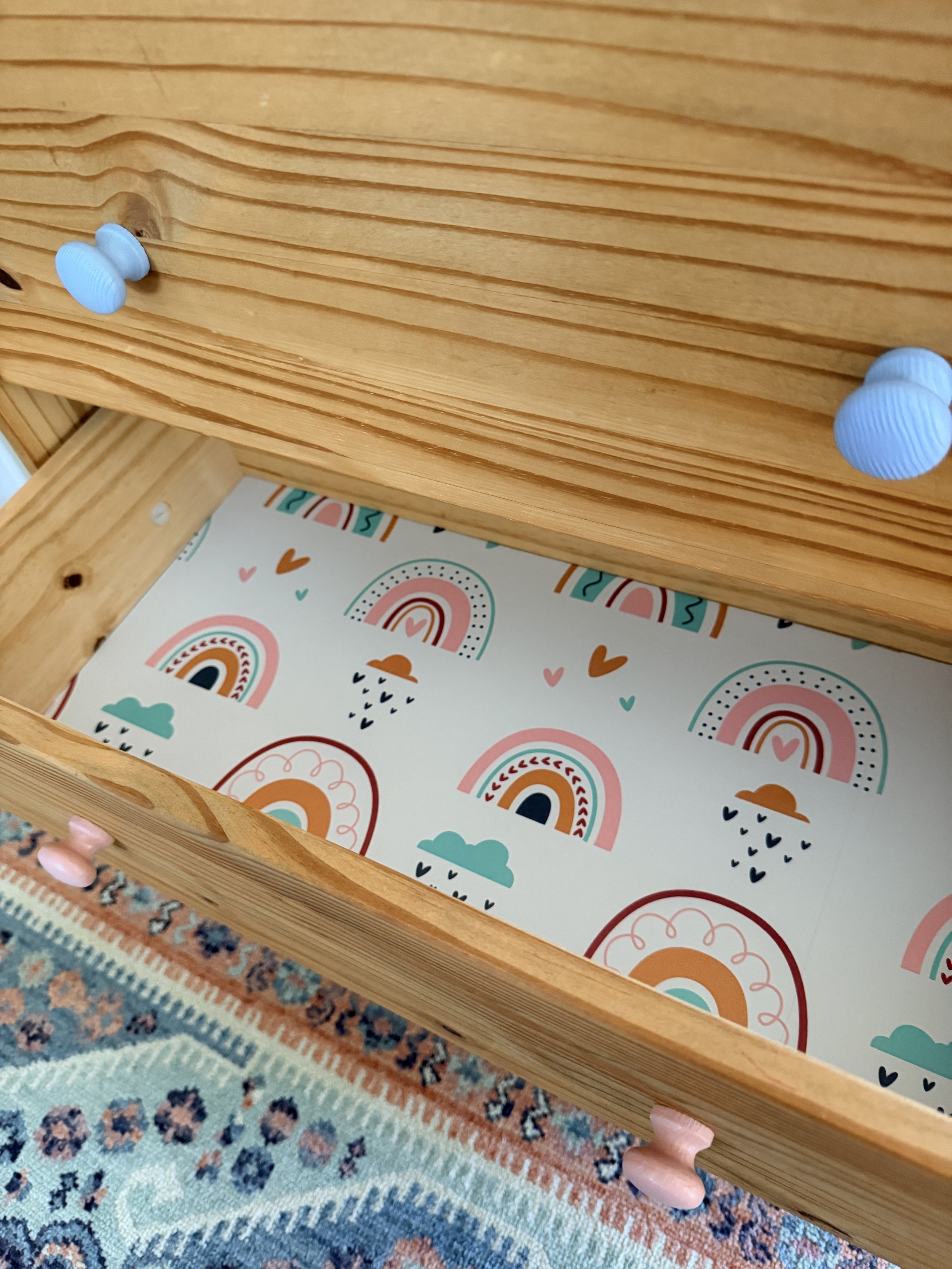 Open wooden drawer with pink, blue, and peach knobs, revealing a colorful rainbow and cloud patterned liner inside.