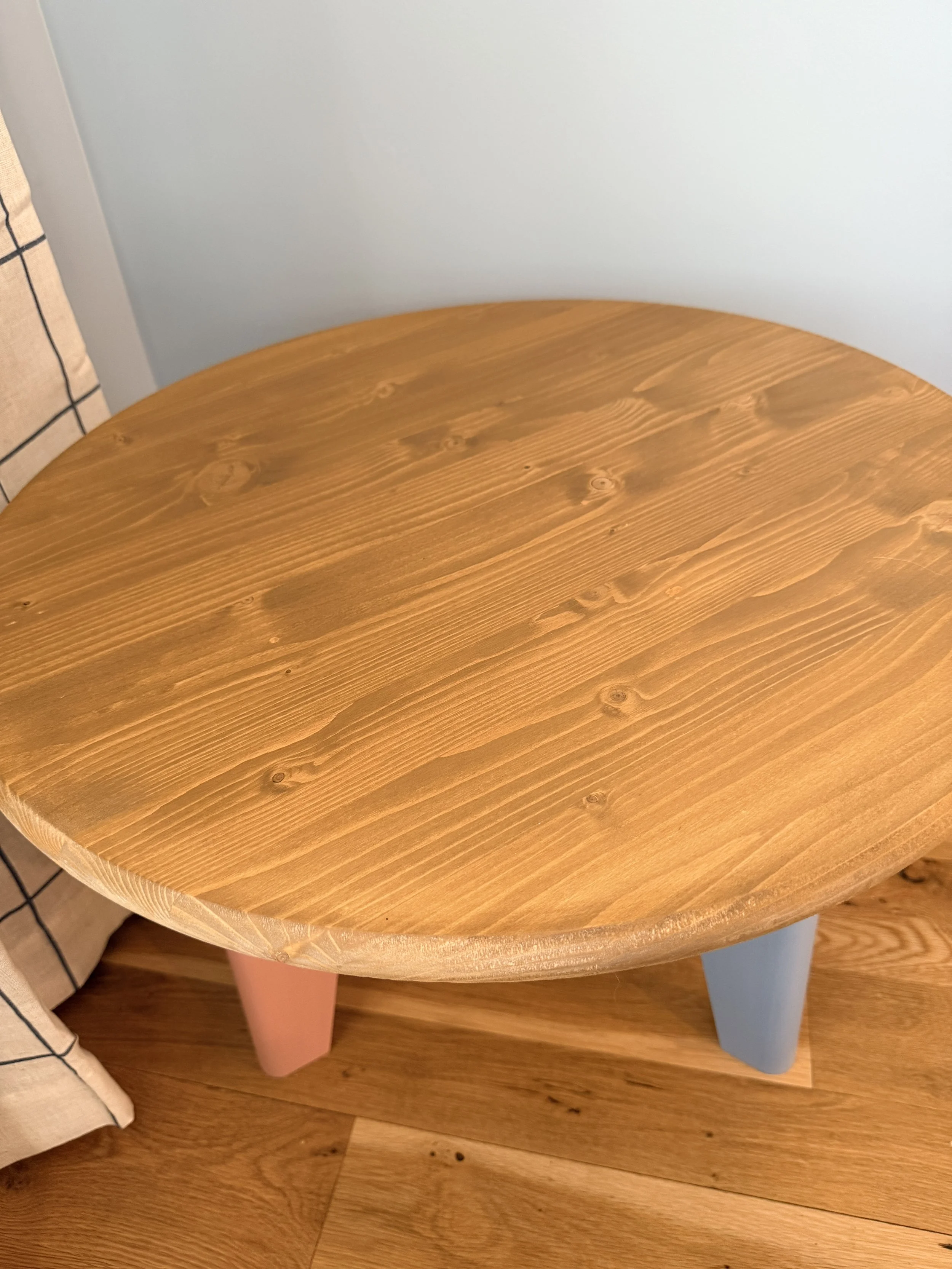 Round wooden coffee table with pastel-colored legs on a hardwood floor beside a beige and black checkered fabric chair.