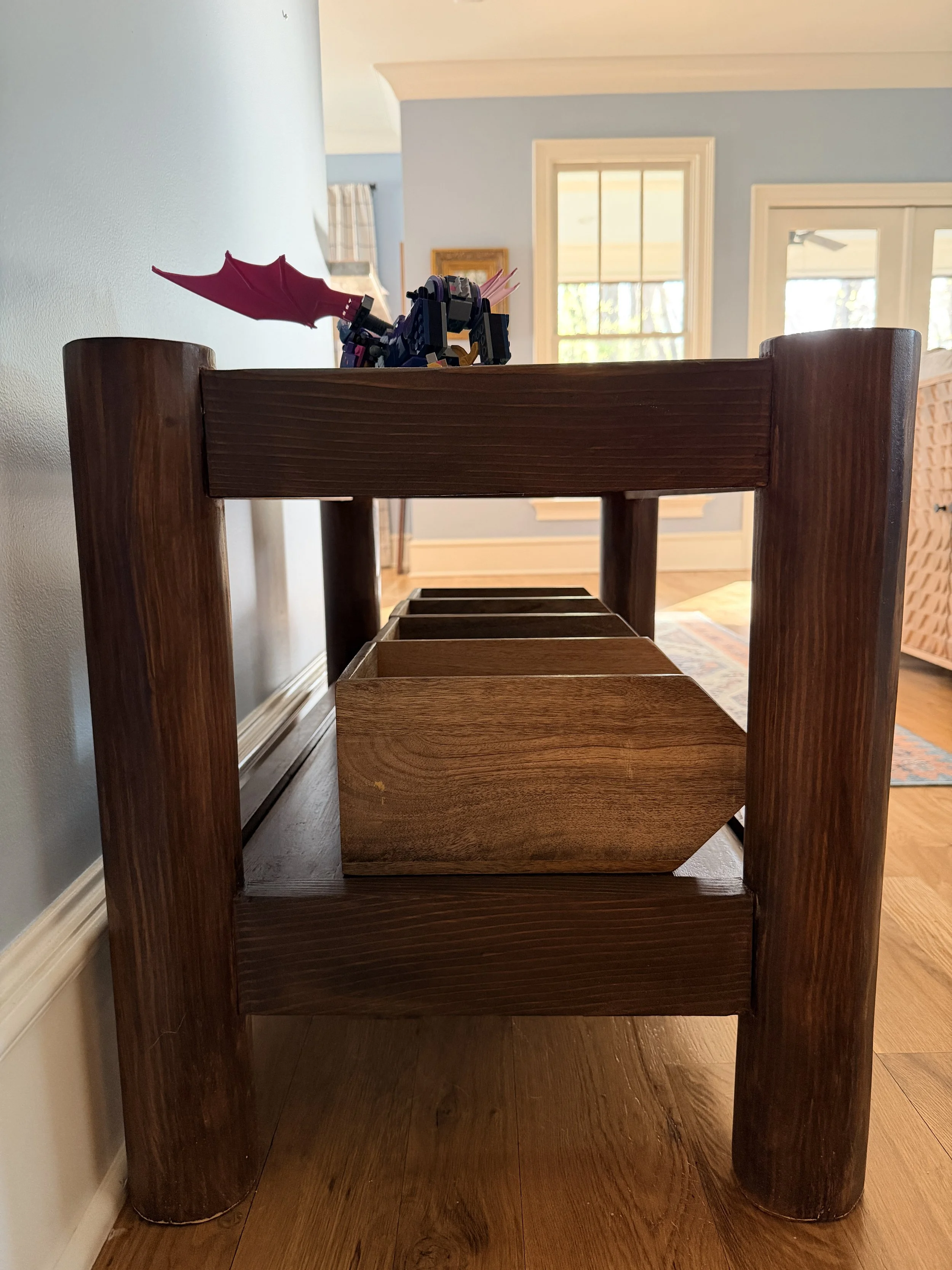 A wooden bookshelf with a toy dragon on top, placed in a room with hardwood floors and light blue walls, near windows and a door.