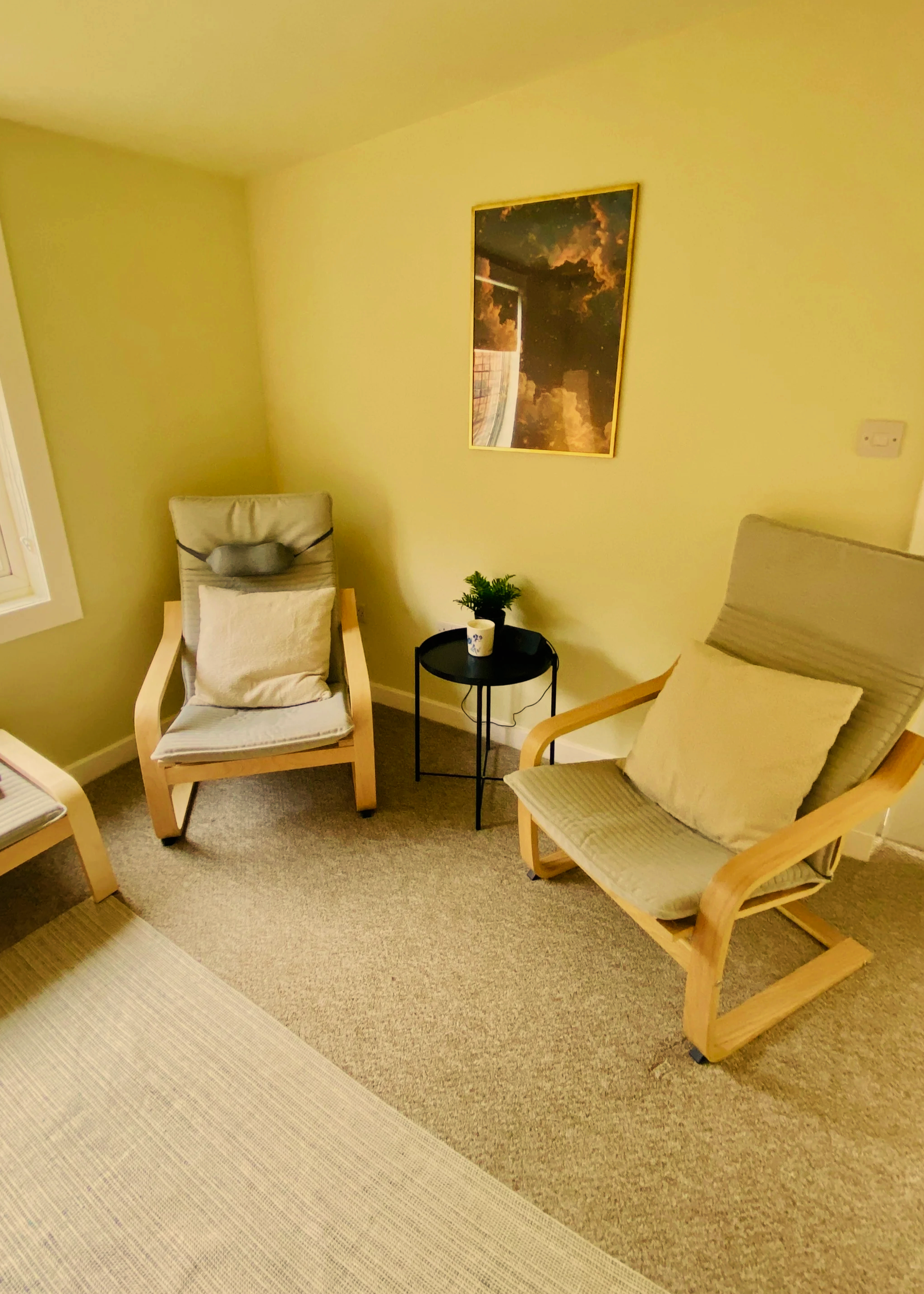 A cozy corner with two wooden framed chairs with cushions, a small round black table with a plant and coffee mug in between, a wall painting, and part of a window with natural light, beige carpet and a yellow wall.