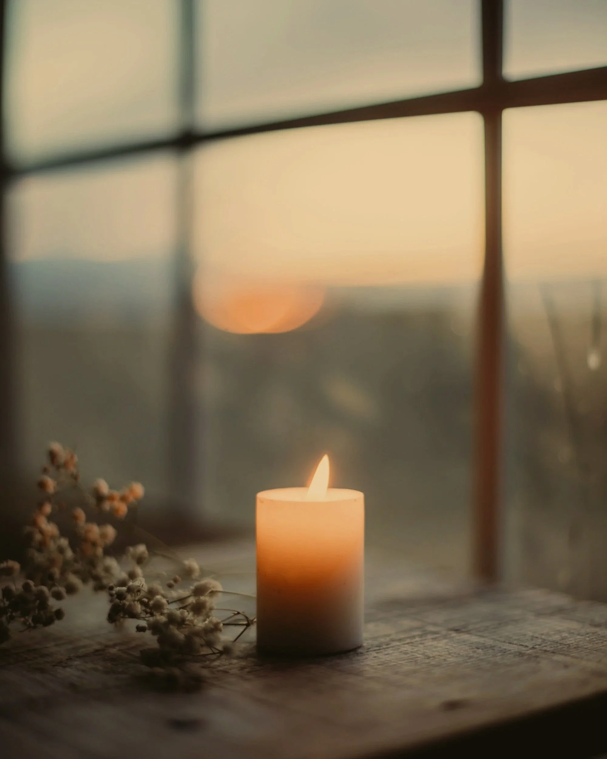 A lit white candle on a wooden surface with a blurred window and sunset in the background.