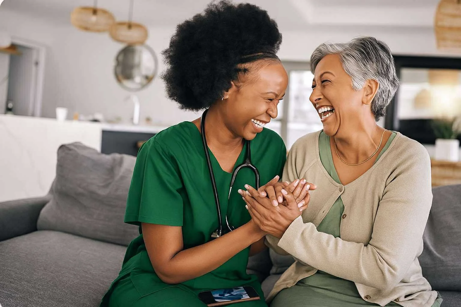 A nurse and an elderly woman laughing and holding hands in a living room.