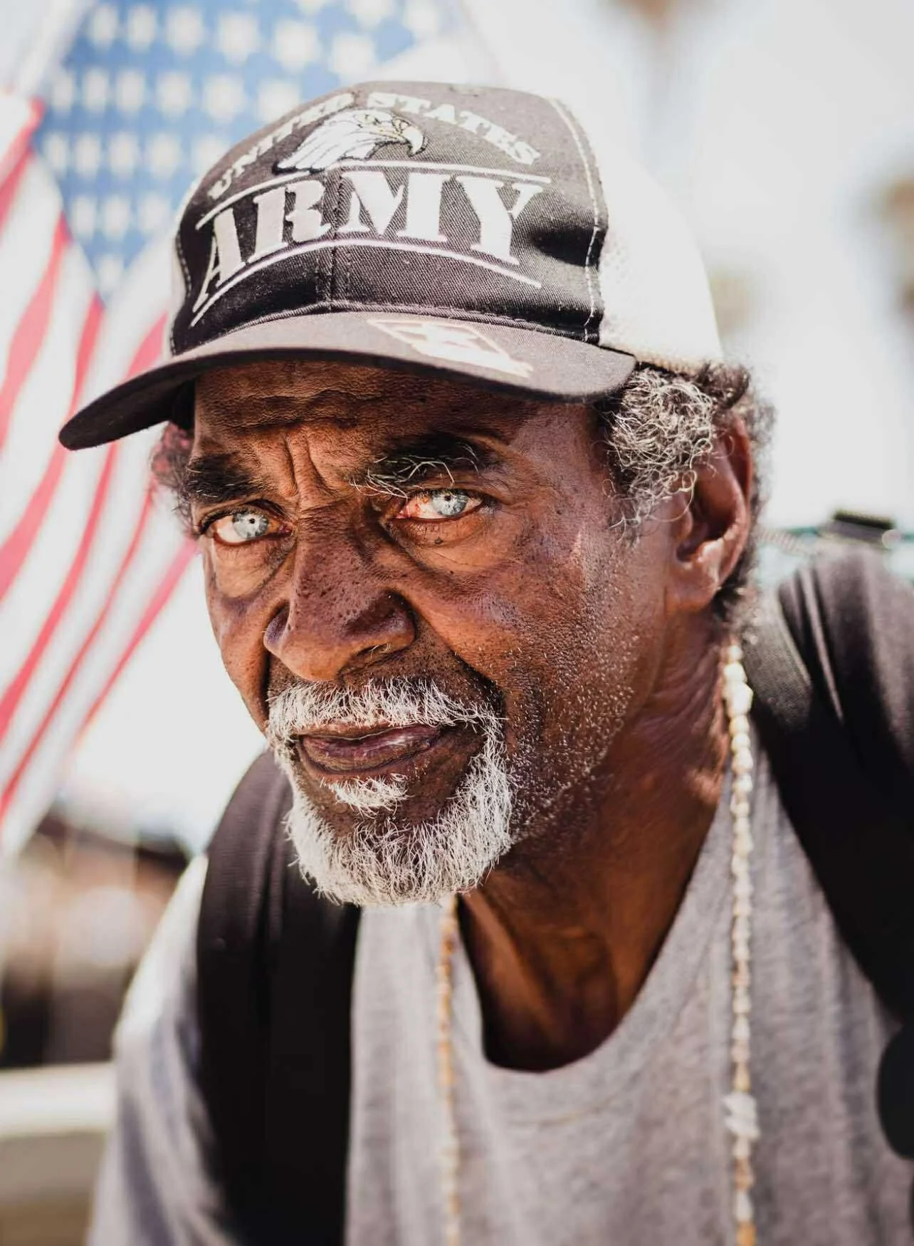Close-up of an elderly man with a white beard and piercing blue eyes, wearing a baseball cap that reads 'U.S. ARMY,' with American flag in the background.