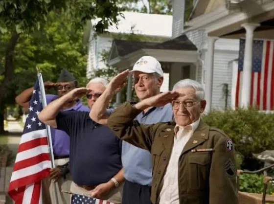 Group of men saluting during a patriotic event, with American flags and a house in the background.