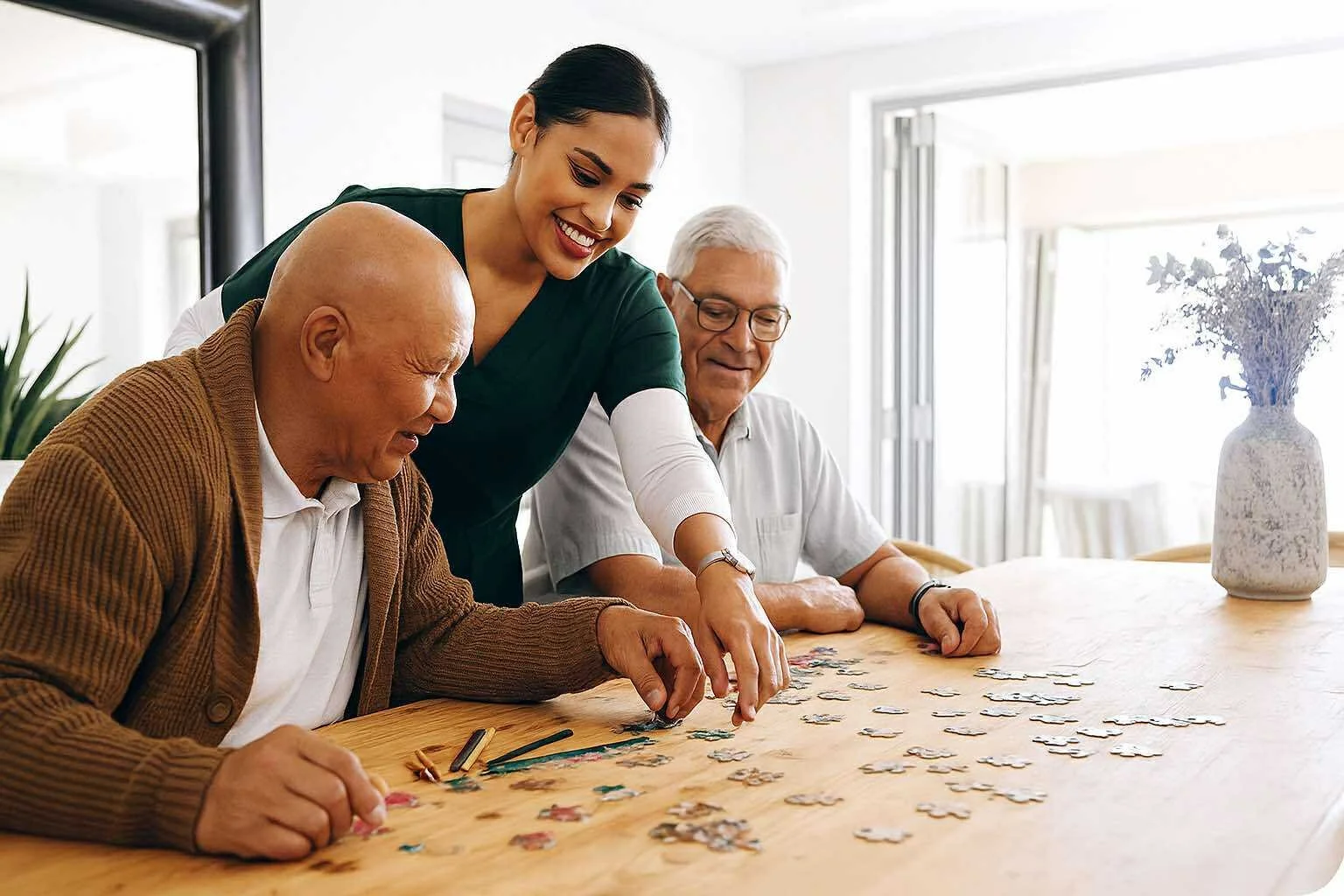 A young woman helps two elderly men assemble a jigsaw puzzle at a wooden table in a bright room with large windows and a vase of dried flowers.