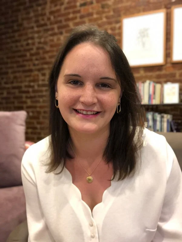 A woman with dark hair, wearing a white blouse, sitting in a room with a brick wall, bookshelves, and framed pictures in the background.