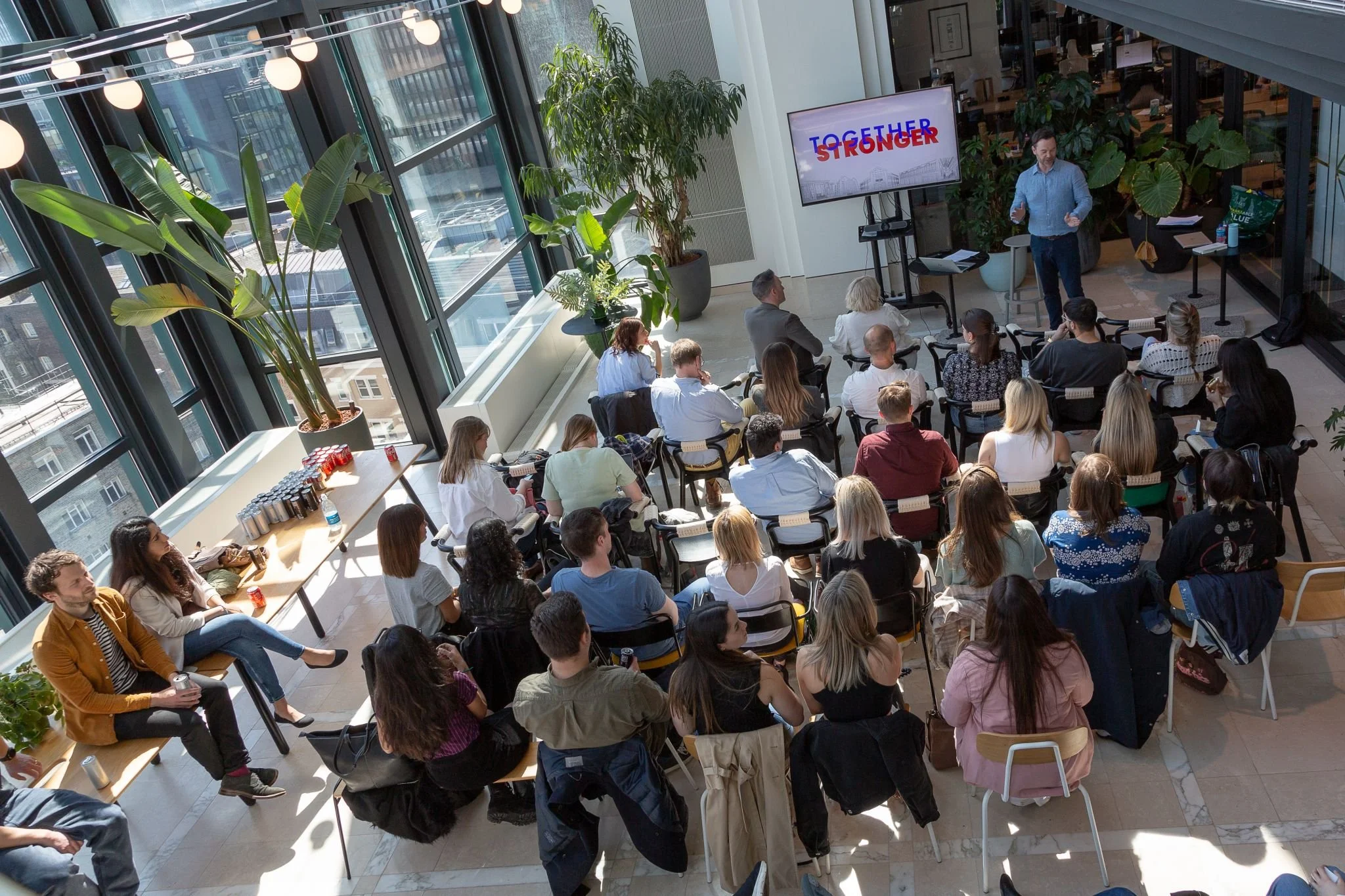 A diverse group of people attending a presentation or seminar in a modern, well-lit conference room with floor-to-ceiling windows, large plants, and a screen displaying the words 'TOGETHER STRONGER'. The presenter isstanding near the screen, addressing the audience.