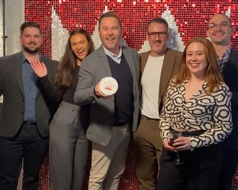 Group of six people including Kevin Murphy celebrating and smiling in front of a heart decoration, with Kevin Murphy holding a Drum Gold award, at a social event or party.