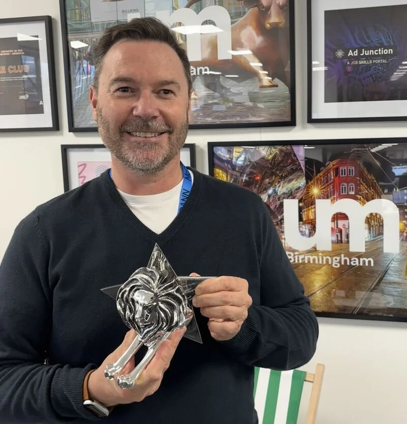 A man named Kevin Murphy with a beard and gray hair smiling and holding a prestigious cannes silver lion-shaped trophy in front of framed pictures on a wall, including one with the text 'UM Birmingham'.