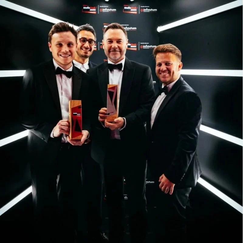 Four men including Kevin Murphy in tuxedos smiling at an awards ceremony, holding tall rectangular prestigious Media Week awards, standing in front of a black background with logos and white diagonal lights.