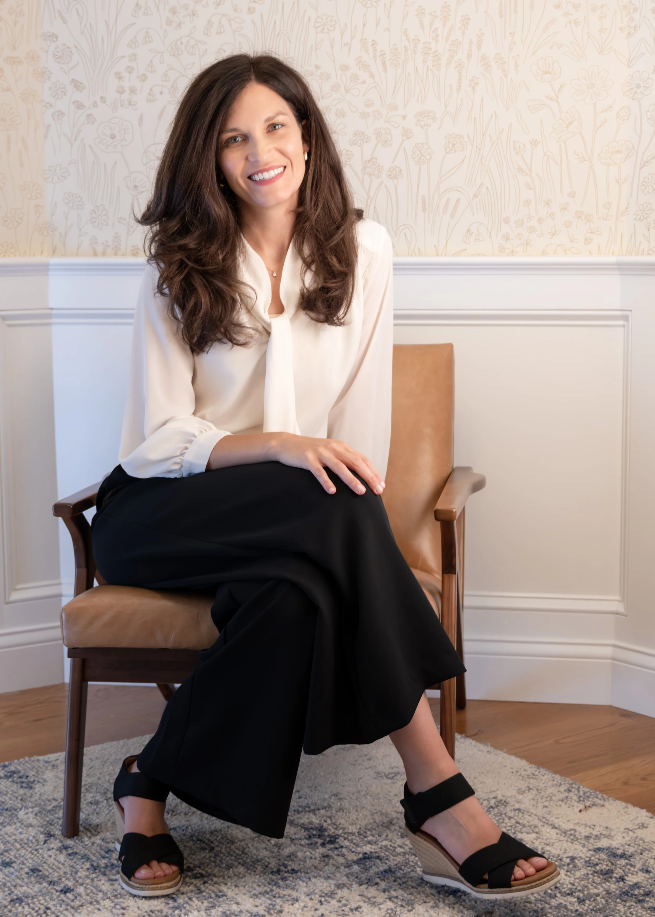 Woman with long dark hair sitting on a chair, smiling, wearing a white blouse, black skirt, and black wedge sandals, in a room with floral wallpaper and a gray patterned carpet.