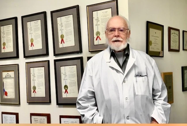 A man in a white lab coat standing in front of a wall with framed diplomas and certificates.