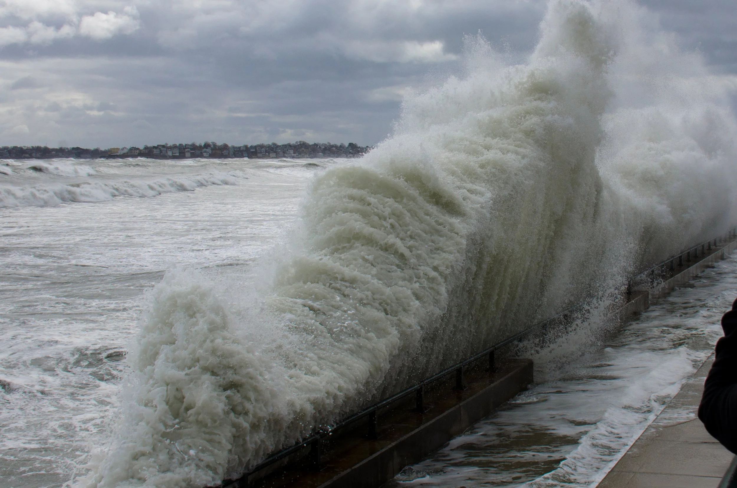 Overcast sky with large ocean waves crashing against the shoreline, with a few buildings visible in the distance and a person standing near the water.