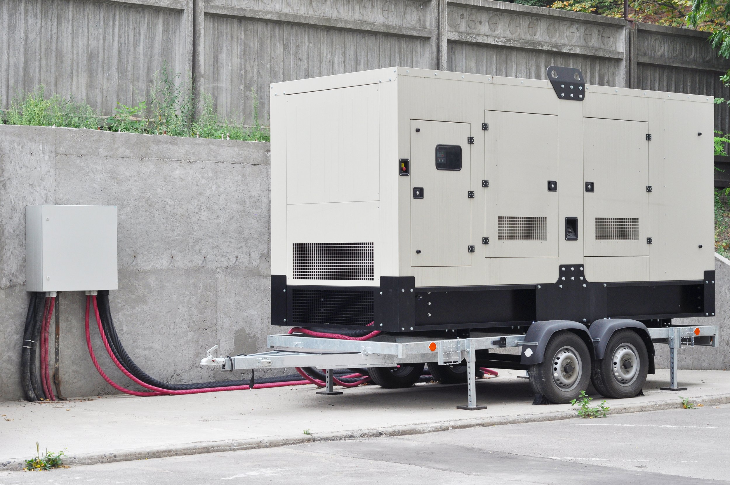 Large industrial generator on trailer parked beside a concrete wall and a small electrical box, with greenery in the background.
