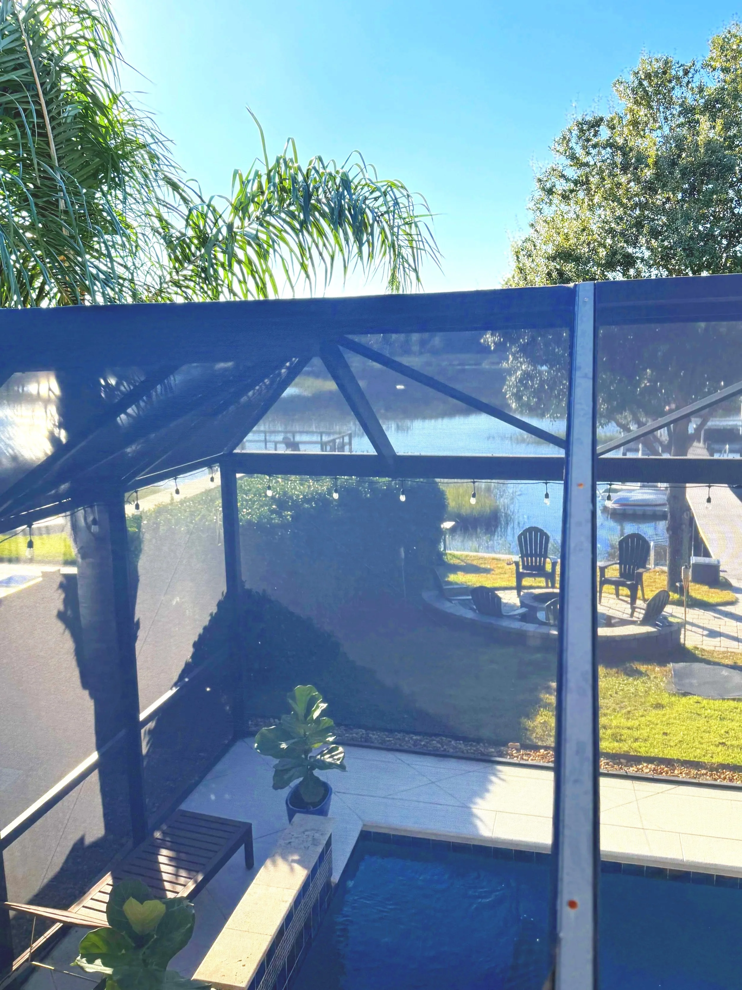 View of a screened-in pool enclosure with an aluminum ladder inside, surrounded by a wooden fence and green trees, on a sunny day.