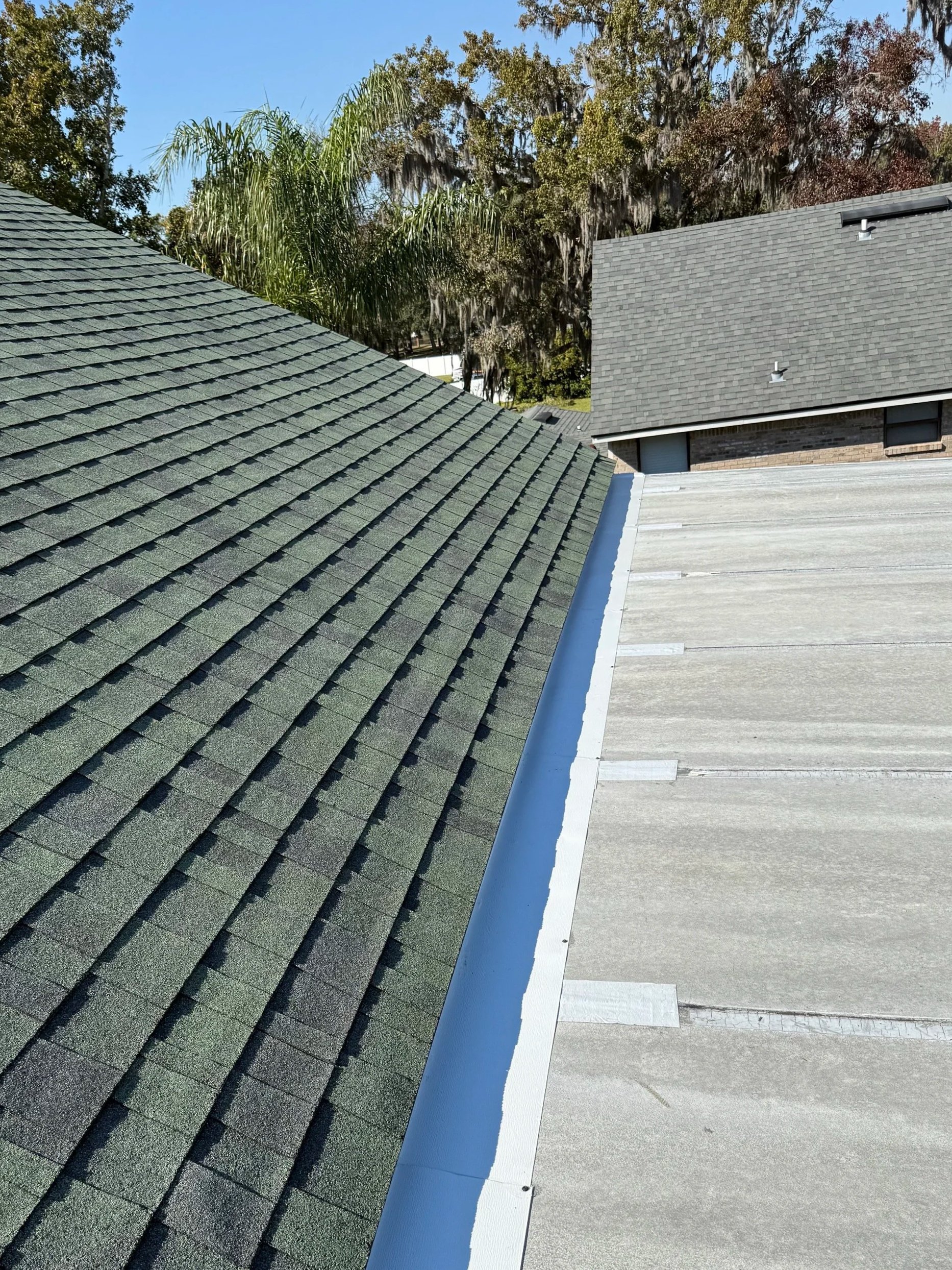 View of a metal roof with weathered patches, a white vinyl fence, green trees, and part of a yellow house in the backyard.