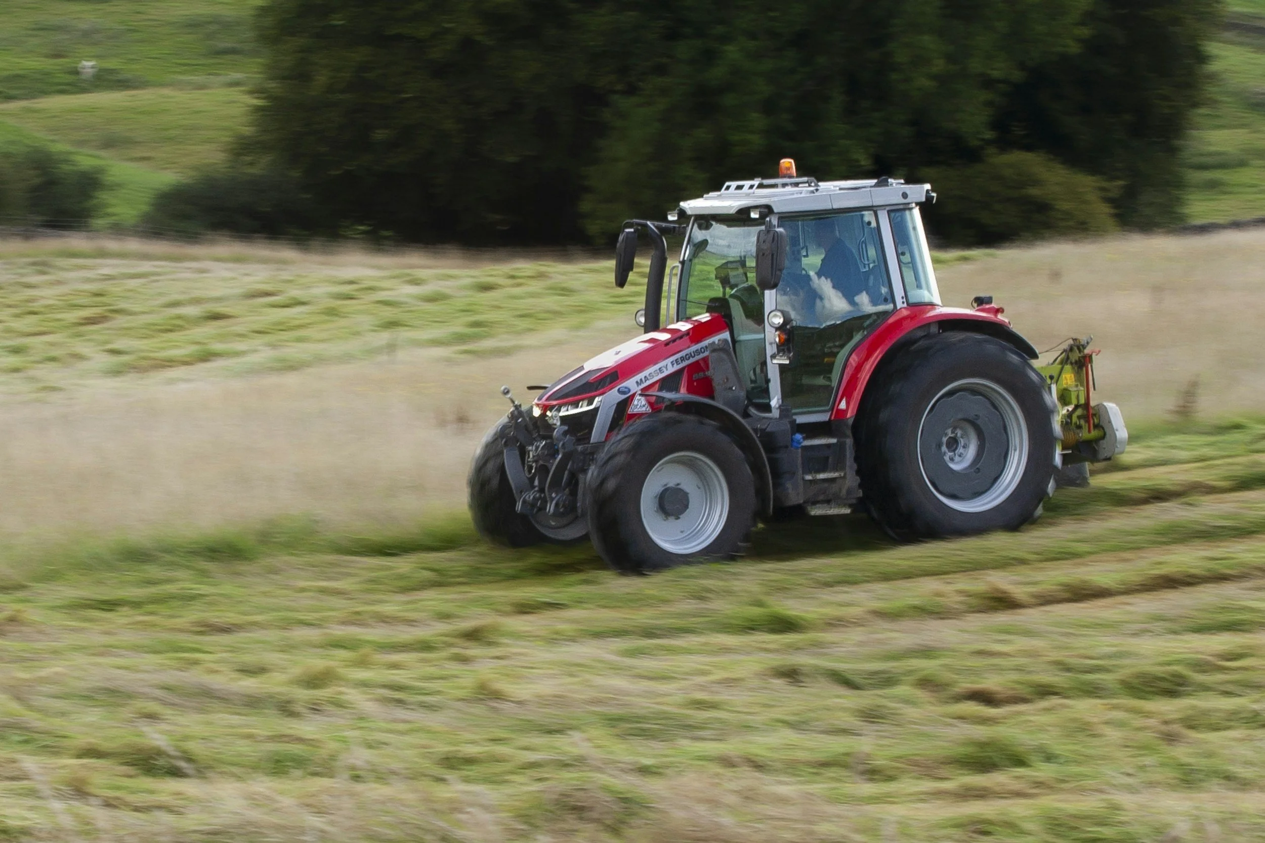 A red tractor is mowing a field of grass on a farm, with trees in the background.