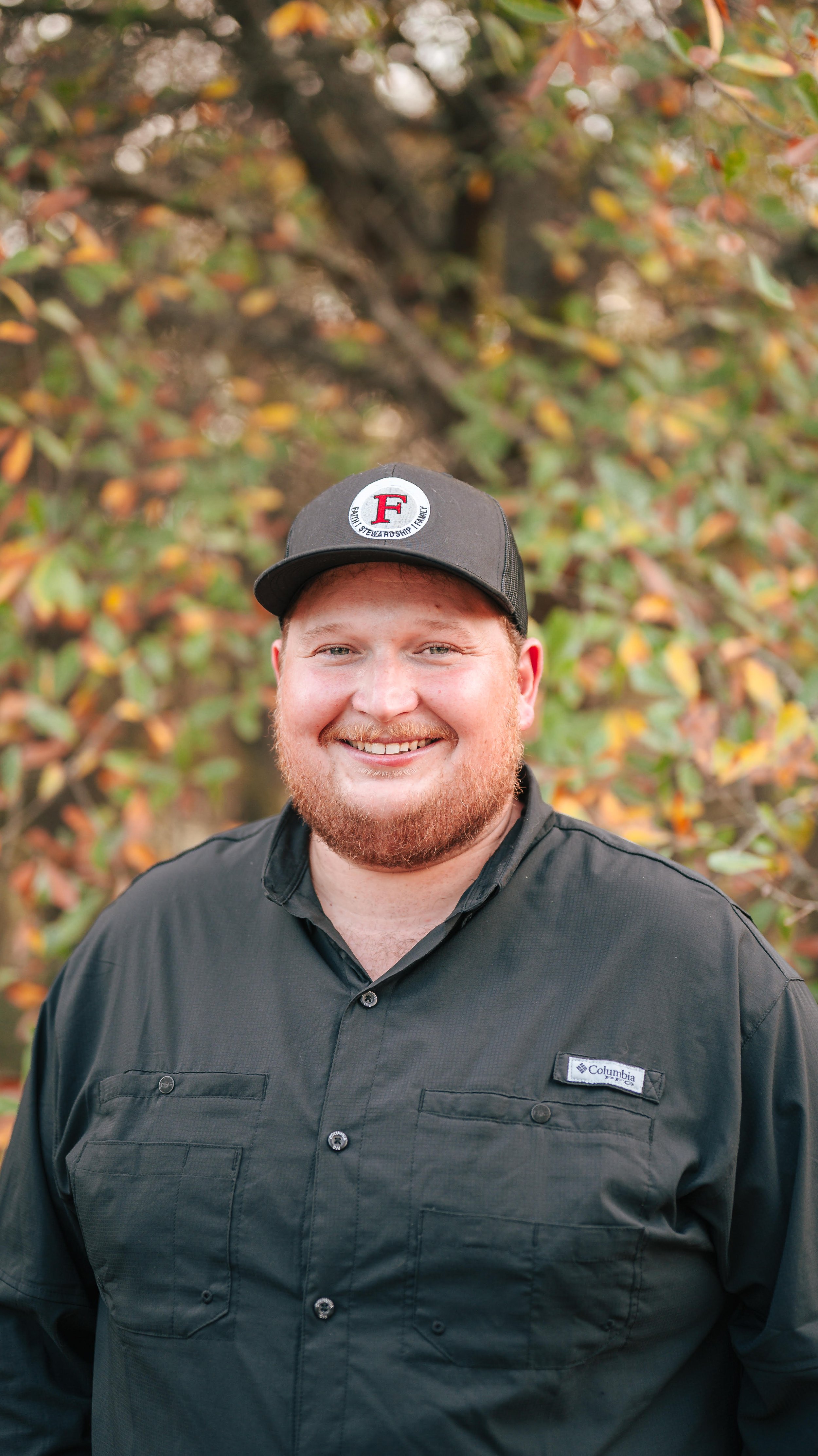 Young man smiling, wearing a camouflage cap with a Syngenta logo, navy Patagonia t-shirt, standing in front of a wooden door and a window with decorative glass, outdoor greenery visible.