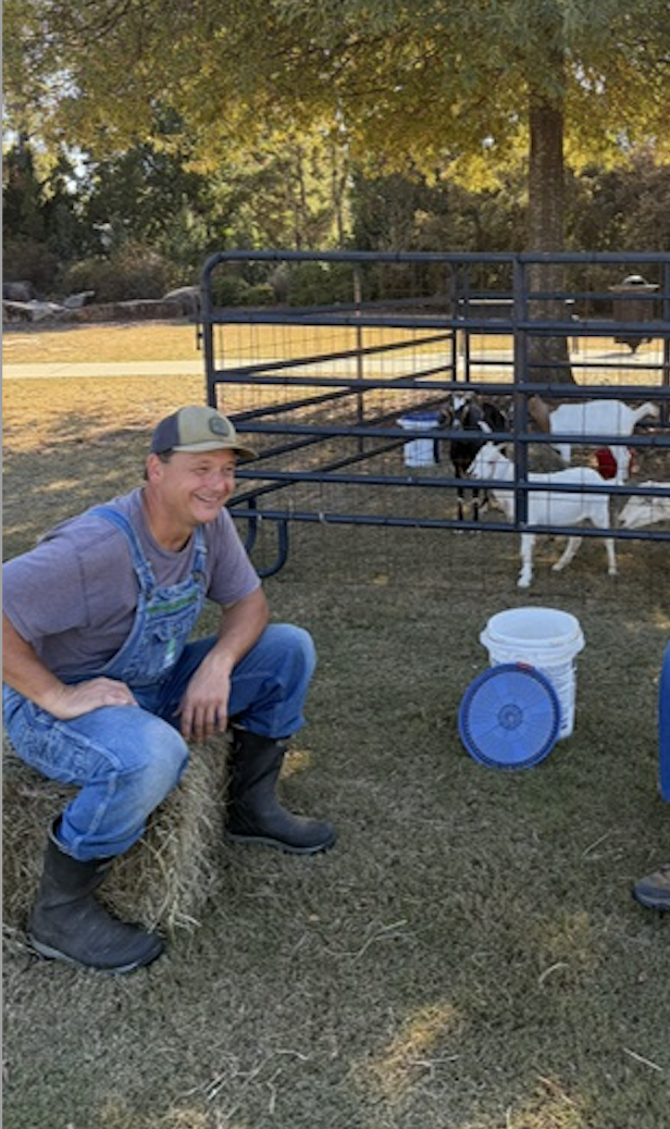 A young man wearing overalls, a gray T-shirt, and a baseball cap sits on a bale of hay, smiling. Behind him is a portable pen with goats of different colors, including white and black, and brown. There are buckets and a fan on the ground.