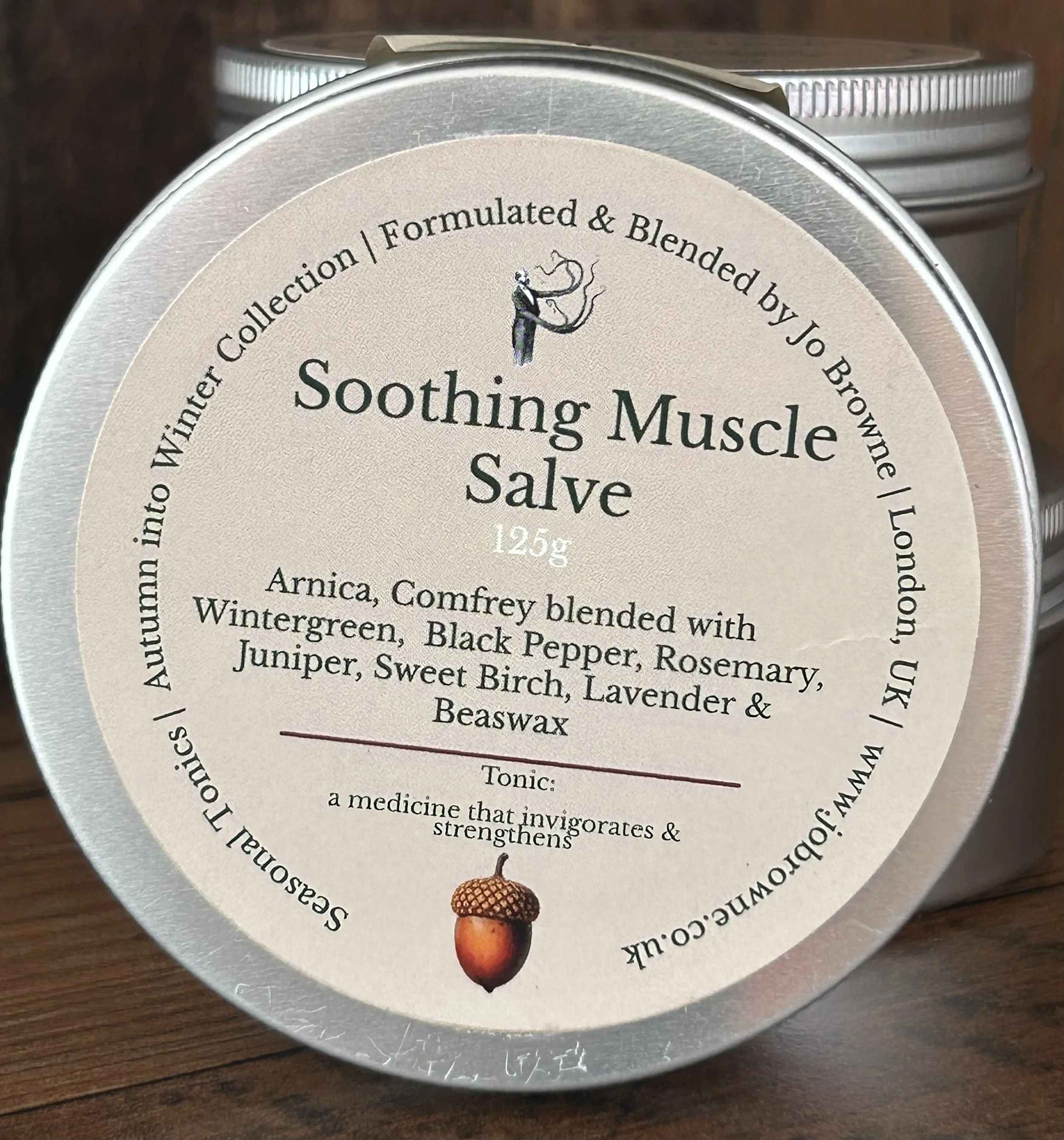 Lid of a round container of Soothing Muscle Salve with a beige label, black text, small illustration of a man smoking, and an acorn image.