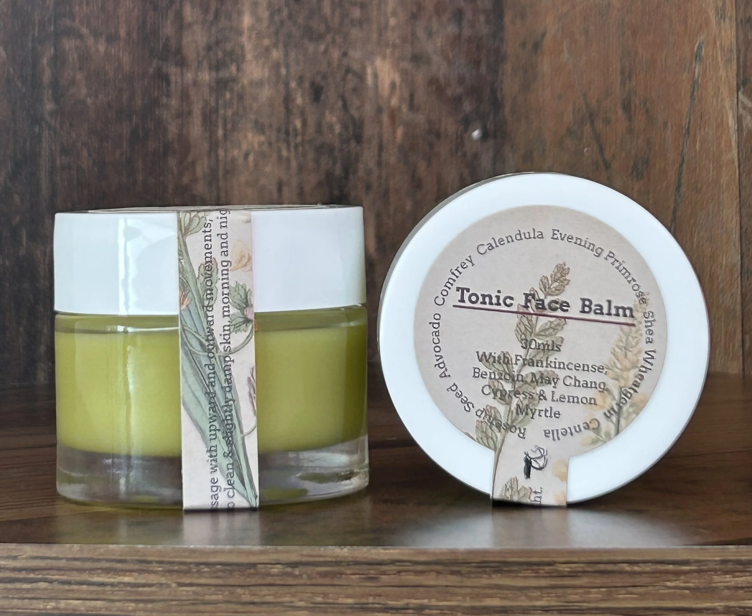 A small glass jar with yellow balm, wrapped with a paper band featuring botanical illustrations, placed next to a round white container labeled 'Tonic Face Balm' with additional product details, on a wooden surface with a dark wood background.
