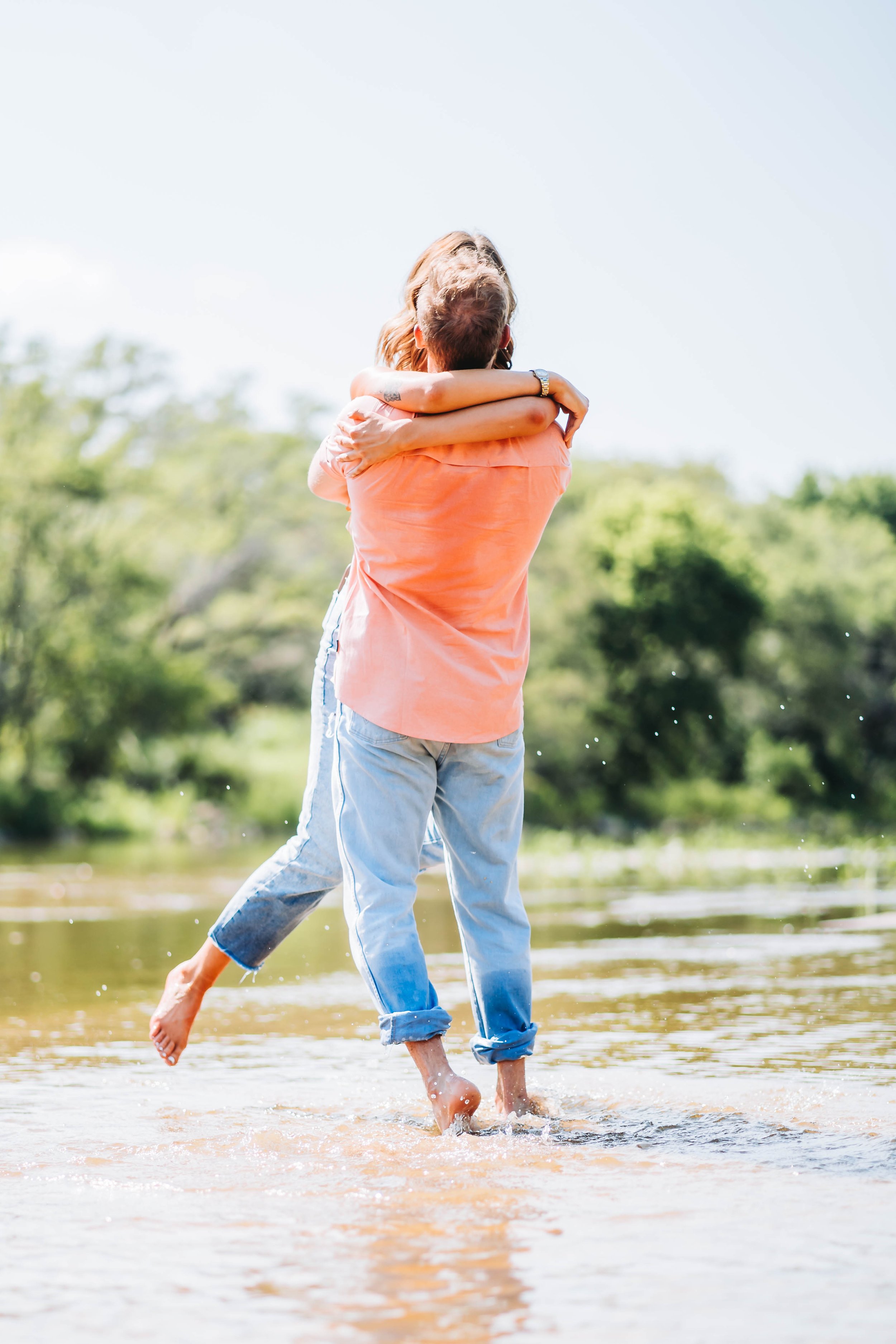 A couple hugging in a shallow river with trees and a bright sky in the background.