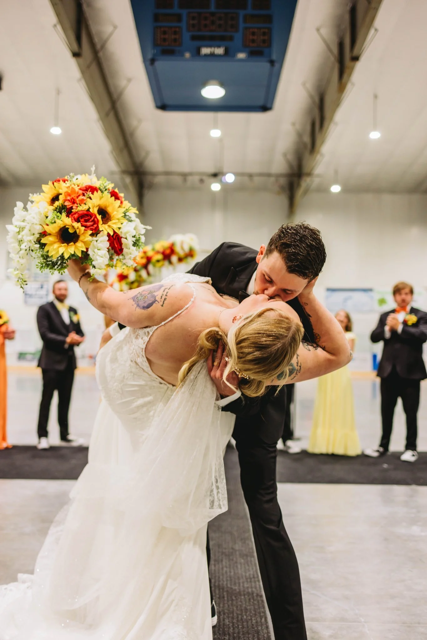 A groom in a black suit kissing a bride in a wedding dress as he dips her during their wedding ceremony, with a bouquet of sunflowers and roses in her hand. Wedding guests are seen in the background clapping.