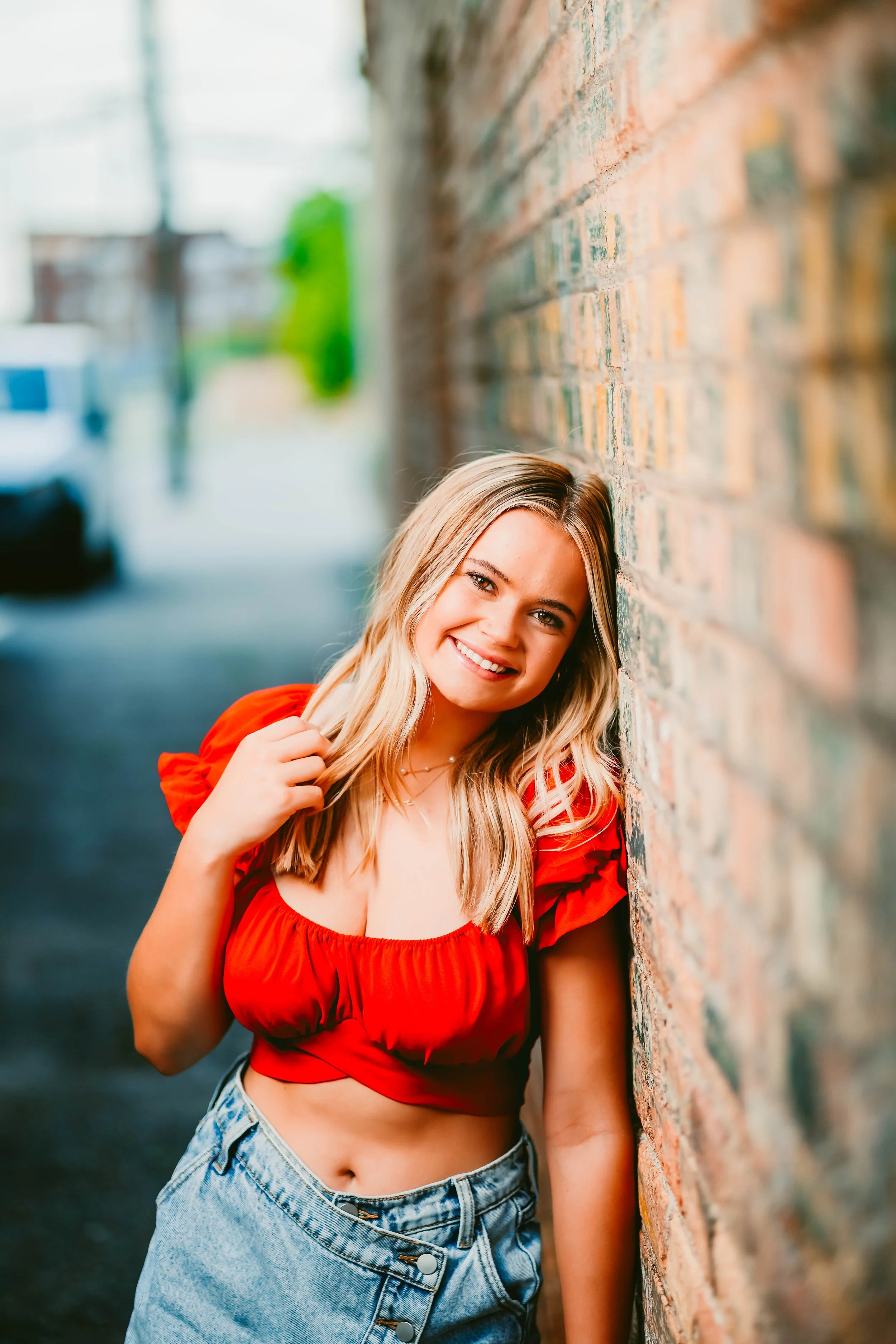 A young woman with blonde hair, wearing a red crop top and denim shorts, leaning against a brick wall outdoors and smiling at the camera.