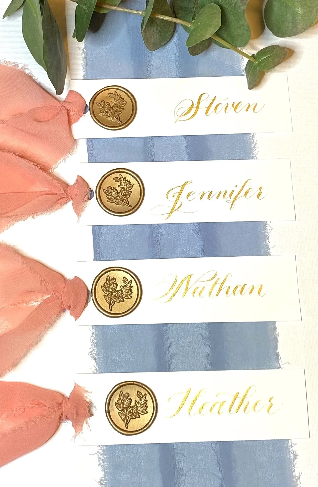 Four white name cards with gold calligraphy and a blue stripe, each decorated with a gold wax seal featuring a floral design, and pink ribbon bows attached to the top left corner of each card. Names on the cards read 'Staven,' 'Jennifer,' 'Mattan,' a