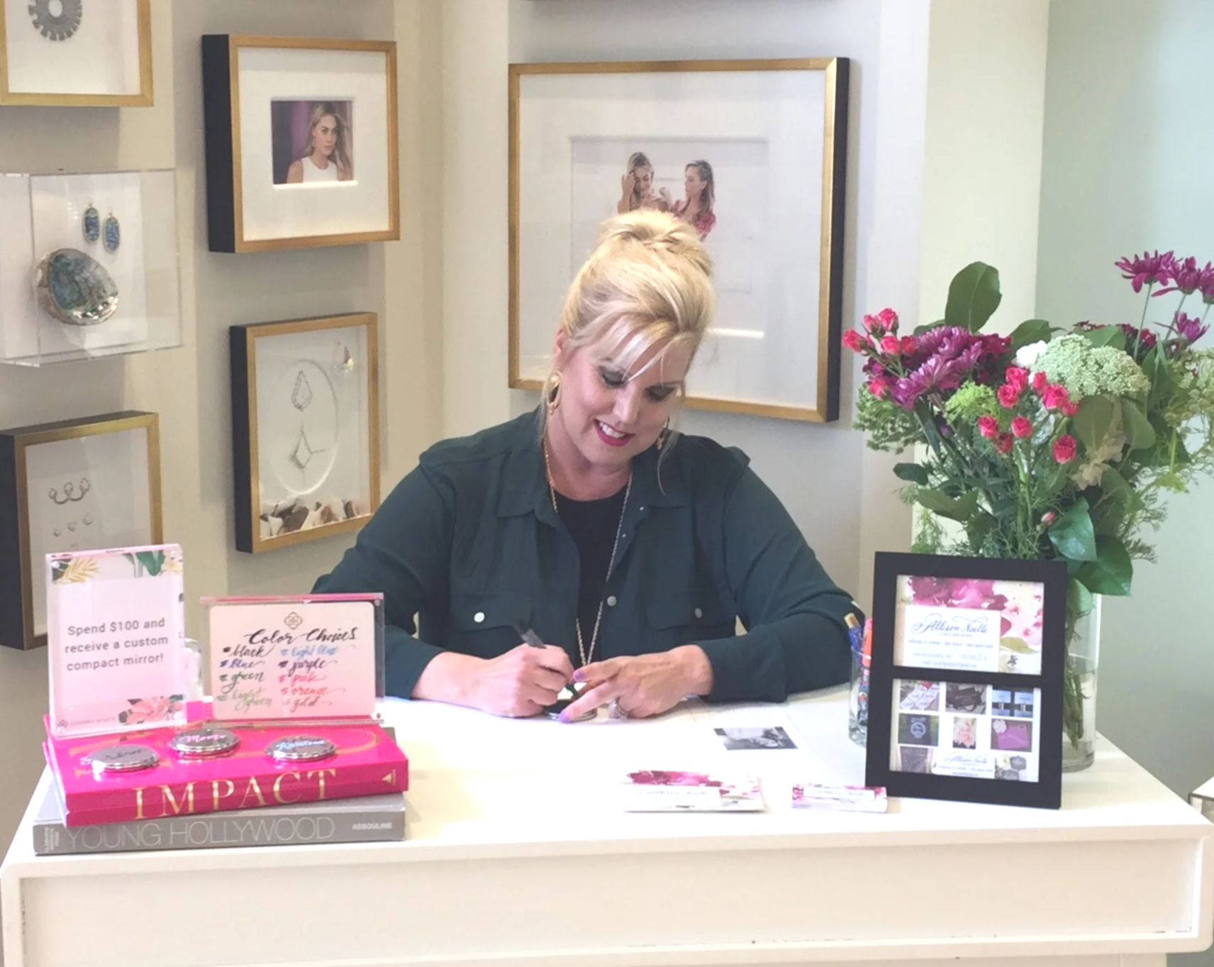 Woman with blonde hair styled in an updo, sitting at a white desk, writing with a pen, surrounded by promotional materials, books, and a large vase of pink and green flowers, with framed pictures on the beige wall behind her.