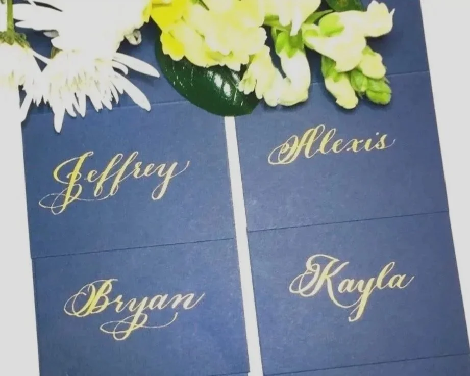 A seating chart with four blue cards featuring gold cursive names: Jeffrey, Bryan, Alexis, and Kayla, with white and yellow flowers at the top.