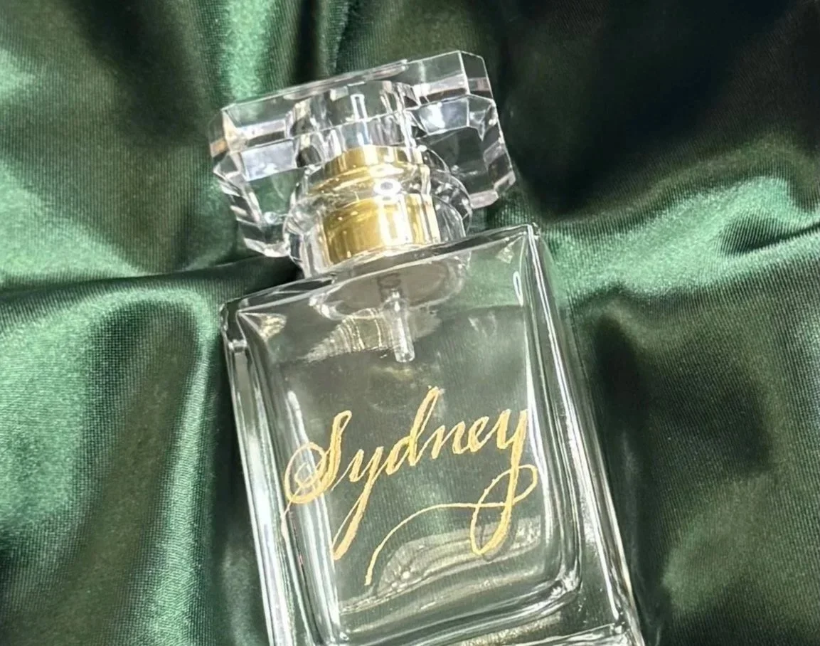 A clear perfume bottle with gold accents and a gold script label reading 'Sydney,' resting on dark green satin fabric.