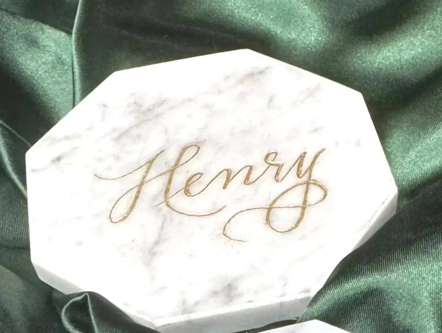 A white marble box with the word 'Henry' written in gold cursive lettering, surrounded by dark green satin fabric.